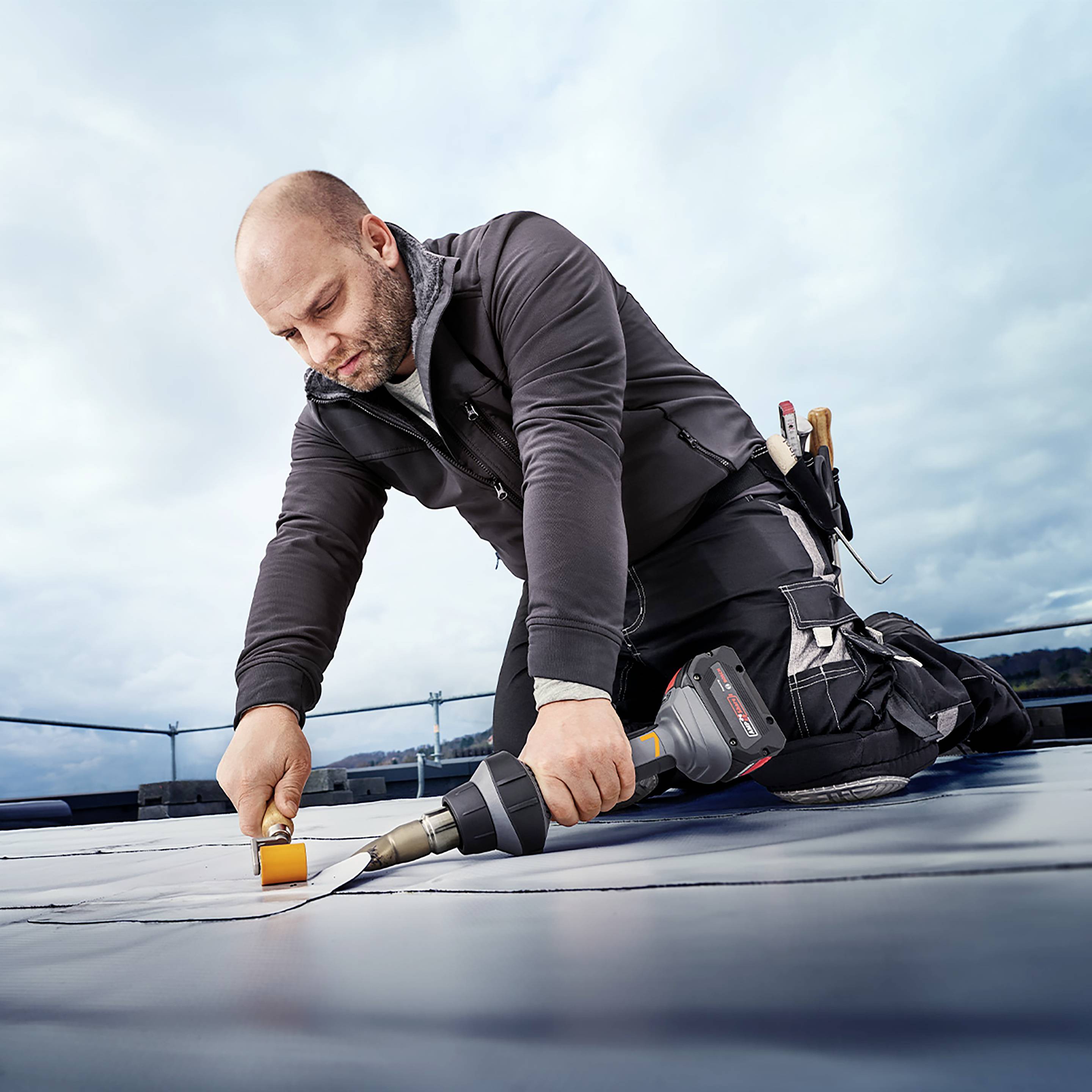 Muškarac radi na ravnom krovu koristeći specijalni alat. Nosi radnu odjeću i koncentriran je na zadatak. U pozadini je nebo oblačno.