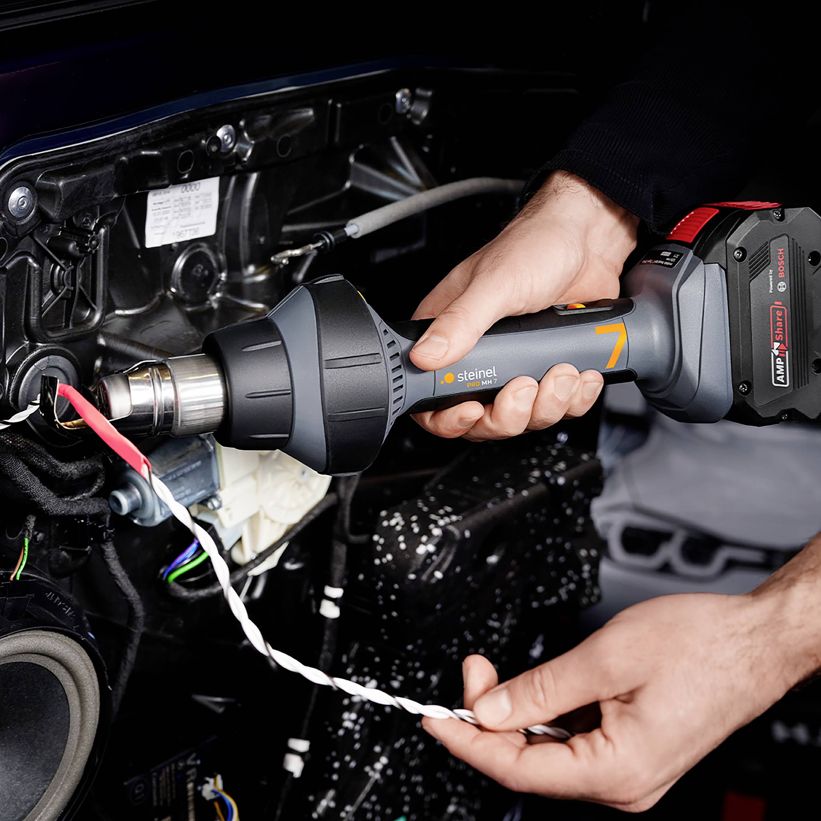 Jedna osoba koristi pištolj vrućeg zraka na tehničkom uređaju. Scena prikazuje precizan rad s naglaskom na detalje.