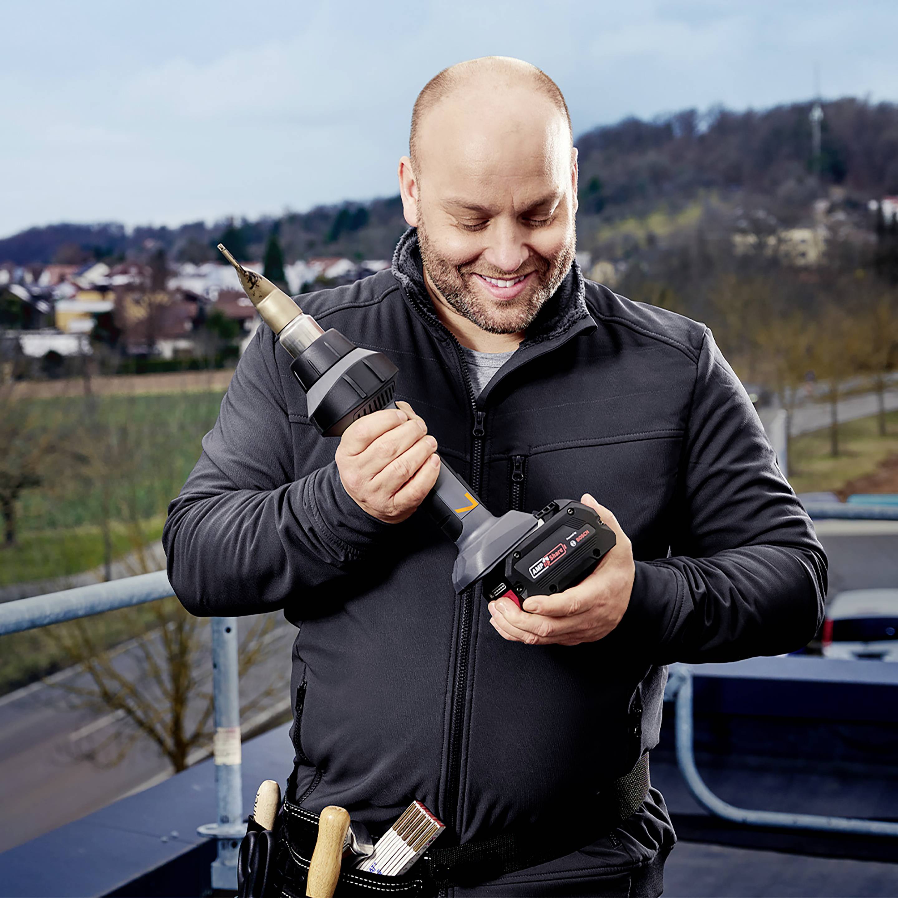 Osoba na balkonu drži akumulatorsku bušilicu i veliki svrdlo, nosi radnu odjeću, smiješi se i promatra alat. U pozadini se prostire gradski krajolik.