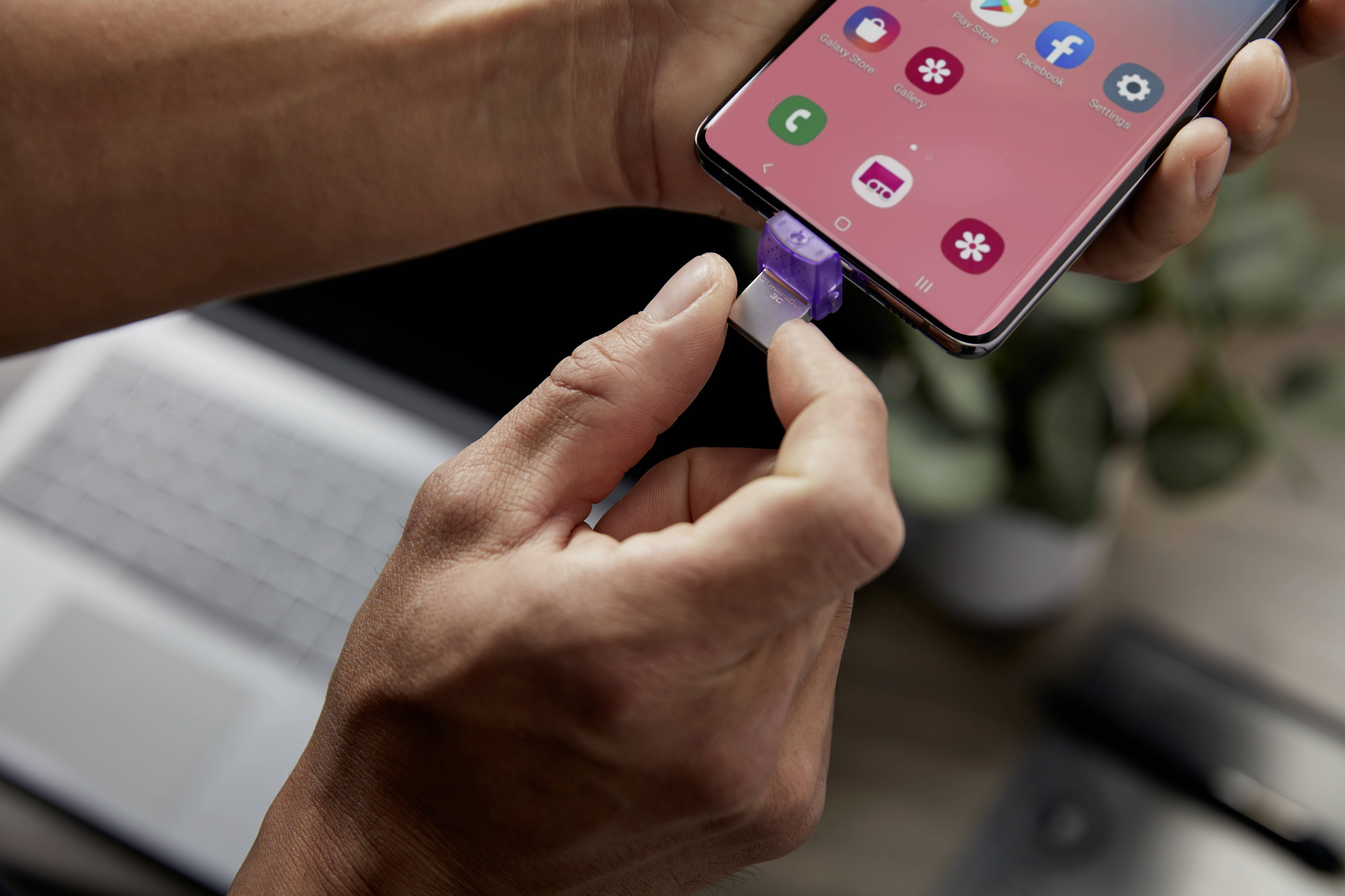 Osoba uključuje punjač u smartphone. Zaslon prikazuje aplikacije. U pozadini se nalaze laptop i biljka.