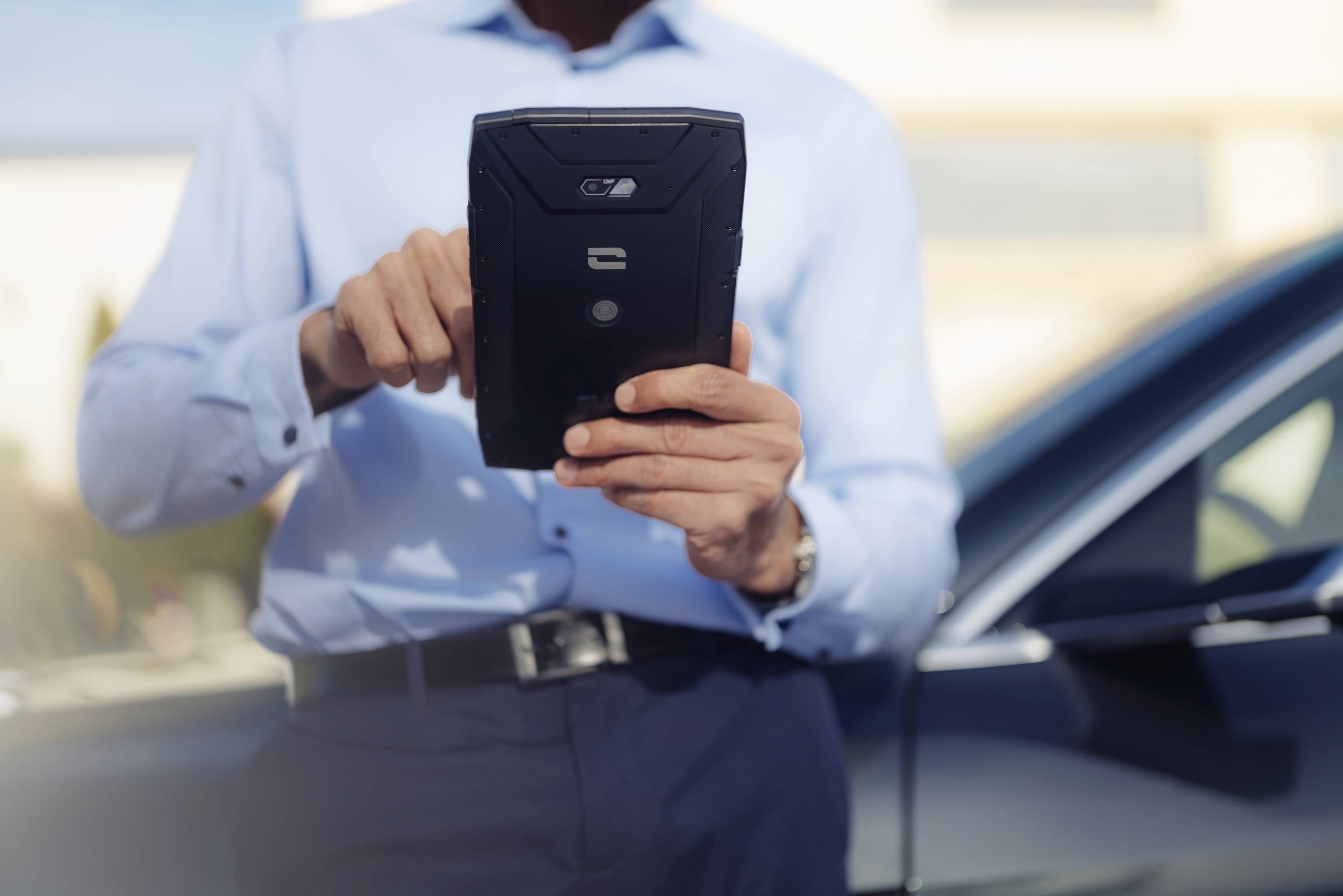 Muškarac u poslovnoj odjeći koristi tablet vani. U pozadini se vidi automobil, što ukazuje na mobilnost ili tehnologiju.