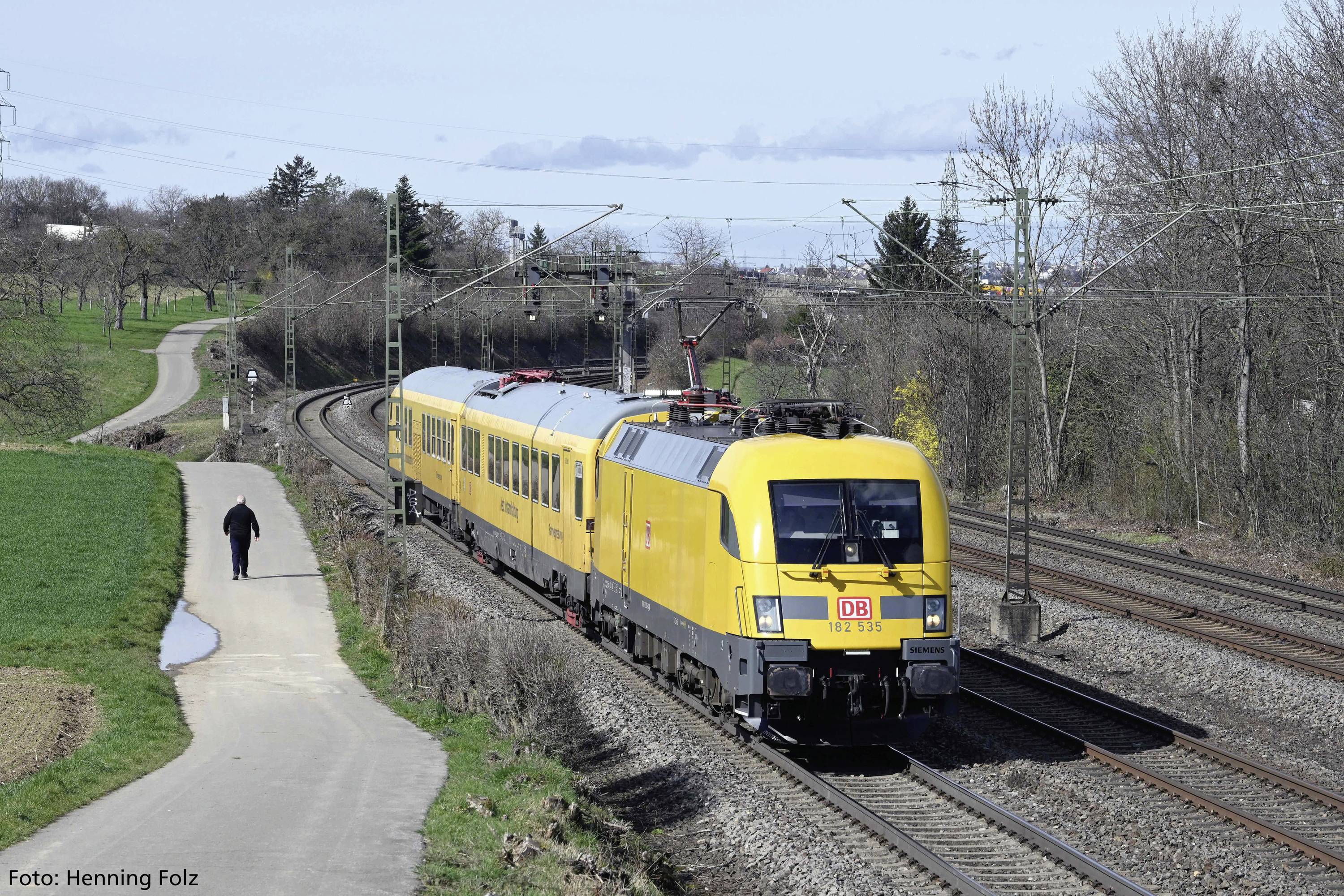 Žuti vlak Deutsche Bahn vozi po dvotračnoj pruzi kroz ruralni krajolik, stabla su s desne strane, čovjek je lijevo.