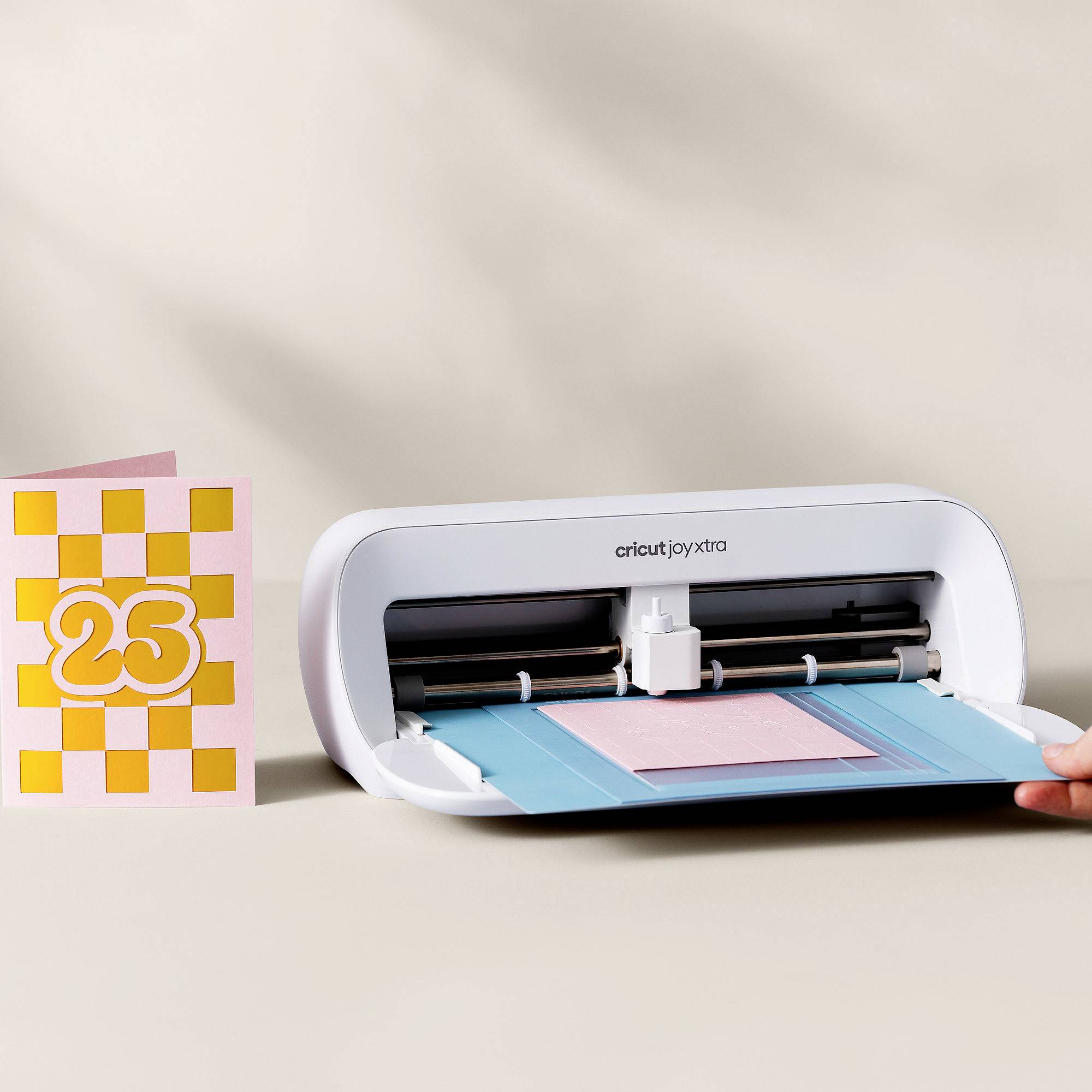 Uređaj Cricut Joy Xtra reže papir. Pored njega se nalazi kartica s brojem '23' u narančastoj i bijeloj šahovskoj kombinaciji.