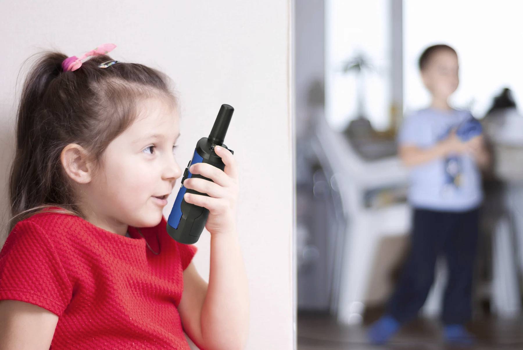 Djevojka u crvenoj haljini govori u walkie-talkie, dok dječak u pozadini drži isti uređaj.