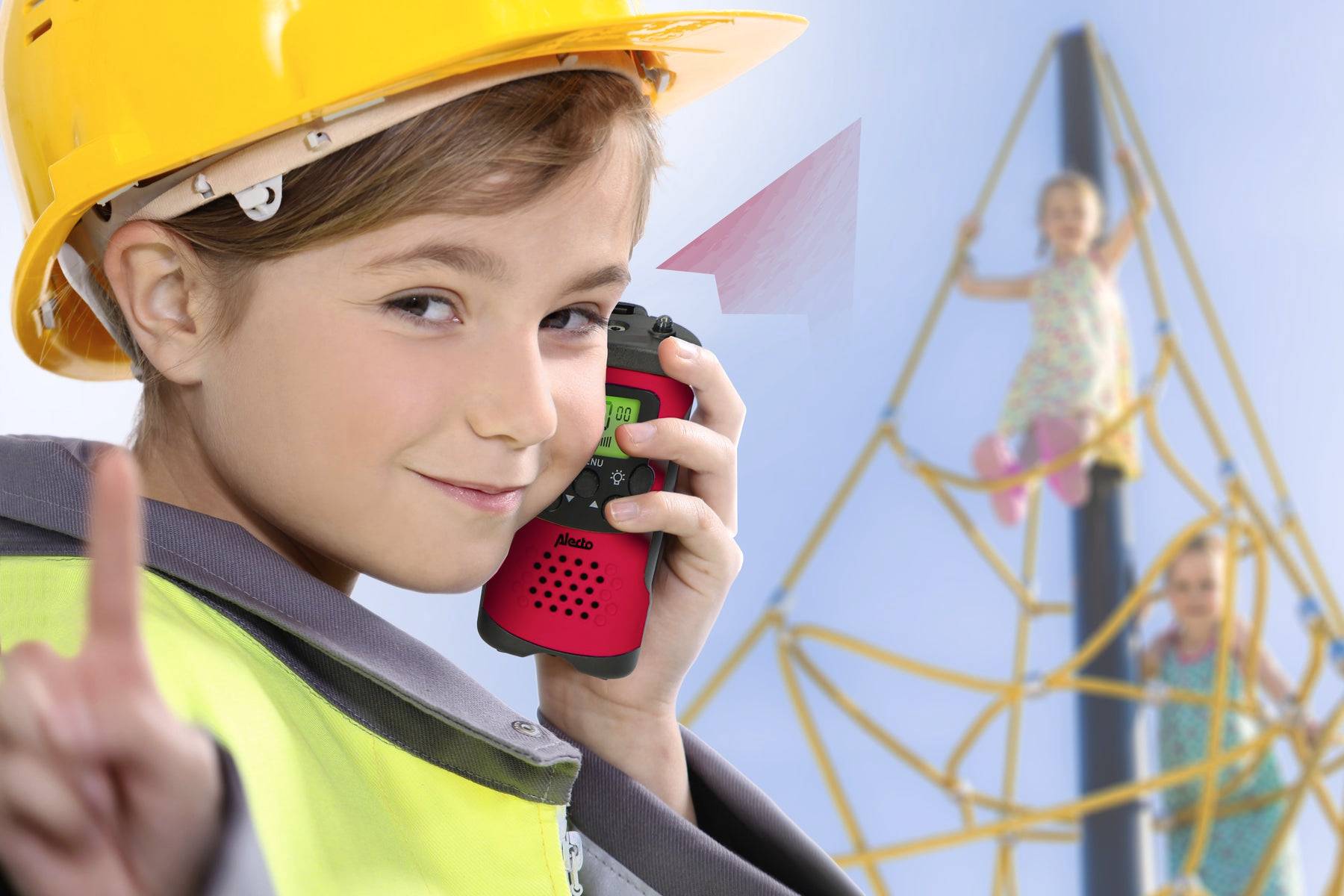 Dijete sa žutom zaštitnom kacigom drži walkie-talkie i smiješi se. U pozadini dvoje djece se igra na penjalištu na dječjem igralištu.