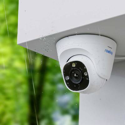 Sigurnosna kamera pričvršćena na zid kuće, prikazuje jasnu leću i infracrvene senzore. Pozadina je zamućena, s drvećem u pozadini.