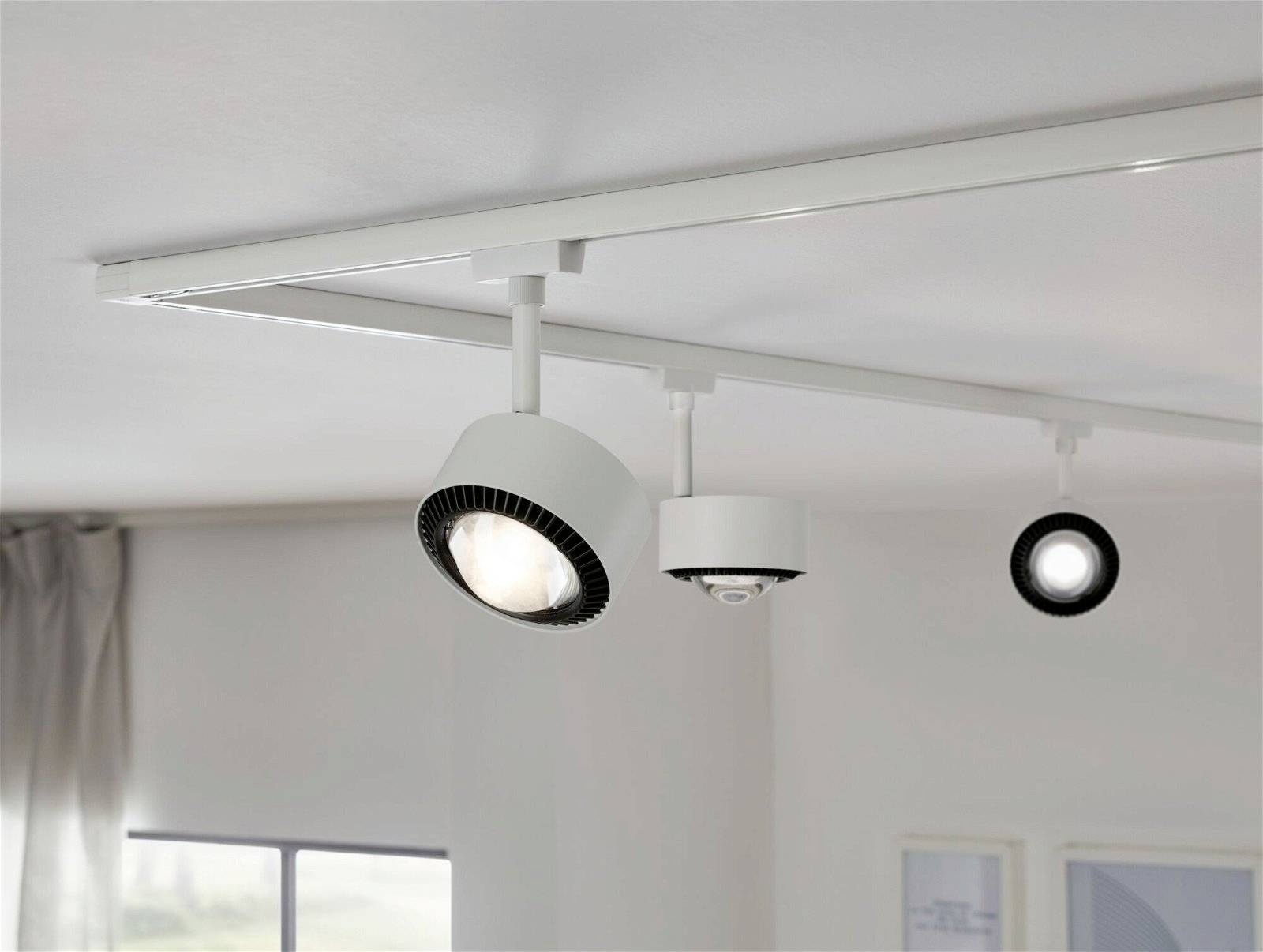 Tri stropne lampe na šini u modernom prostoru. Jedna lampa je uključena. U pozadini se nazire prozorski otvor.