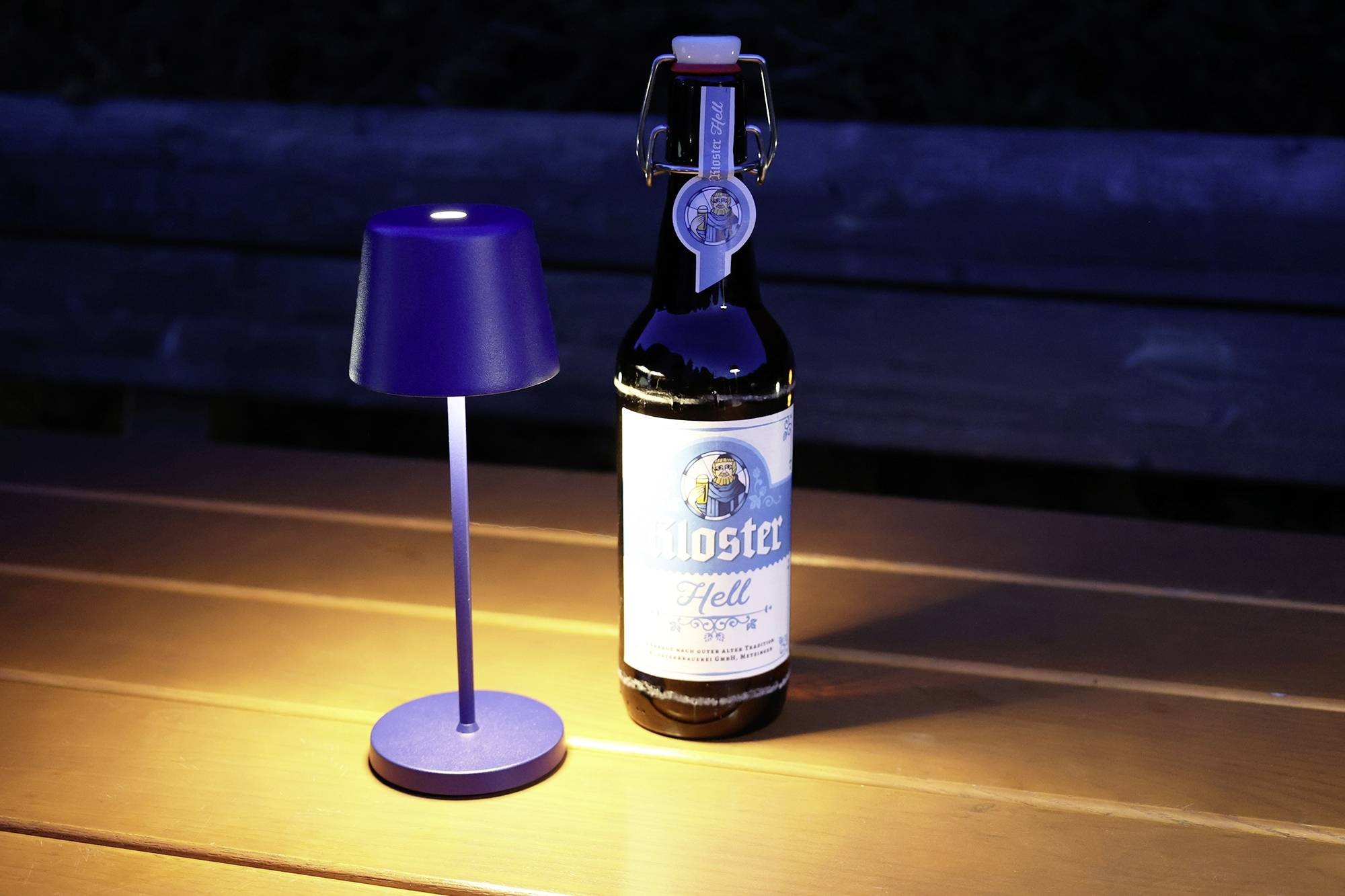 Jedna boca piva s natpisom 'Kloster Hell' stoji na drvenom stolu, pored male, sjajne stolne lampe.