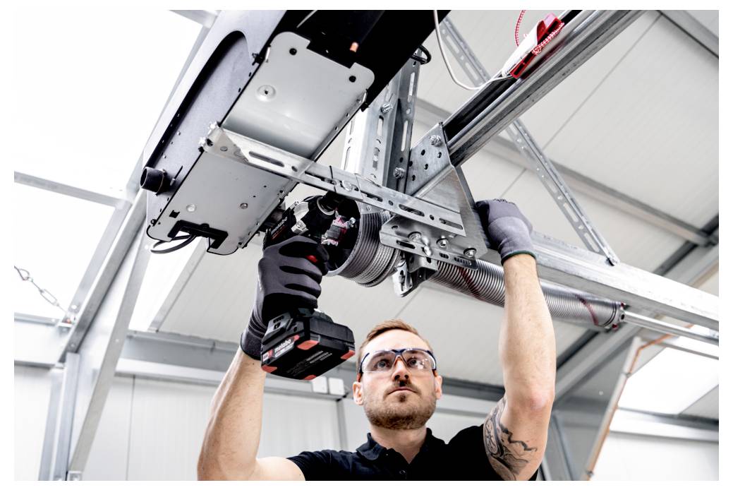 Tehničar koji nosi zaštitne naočale i rukavice koristi ručnu bušilicu za instalaciju ili popravak mehanizma industrijske garažne vrata montiranih na strop.