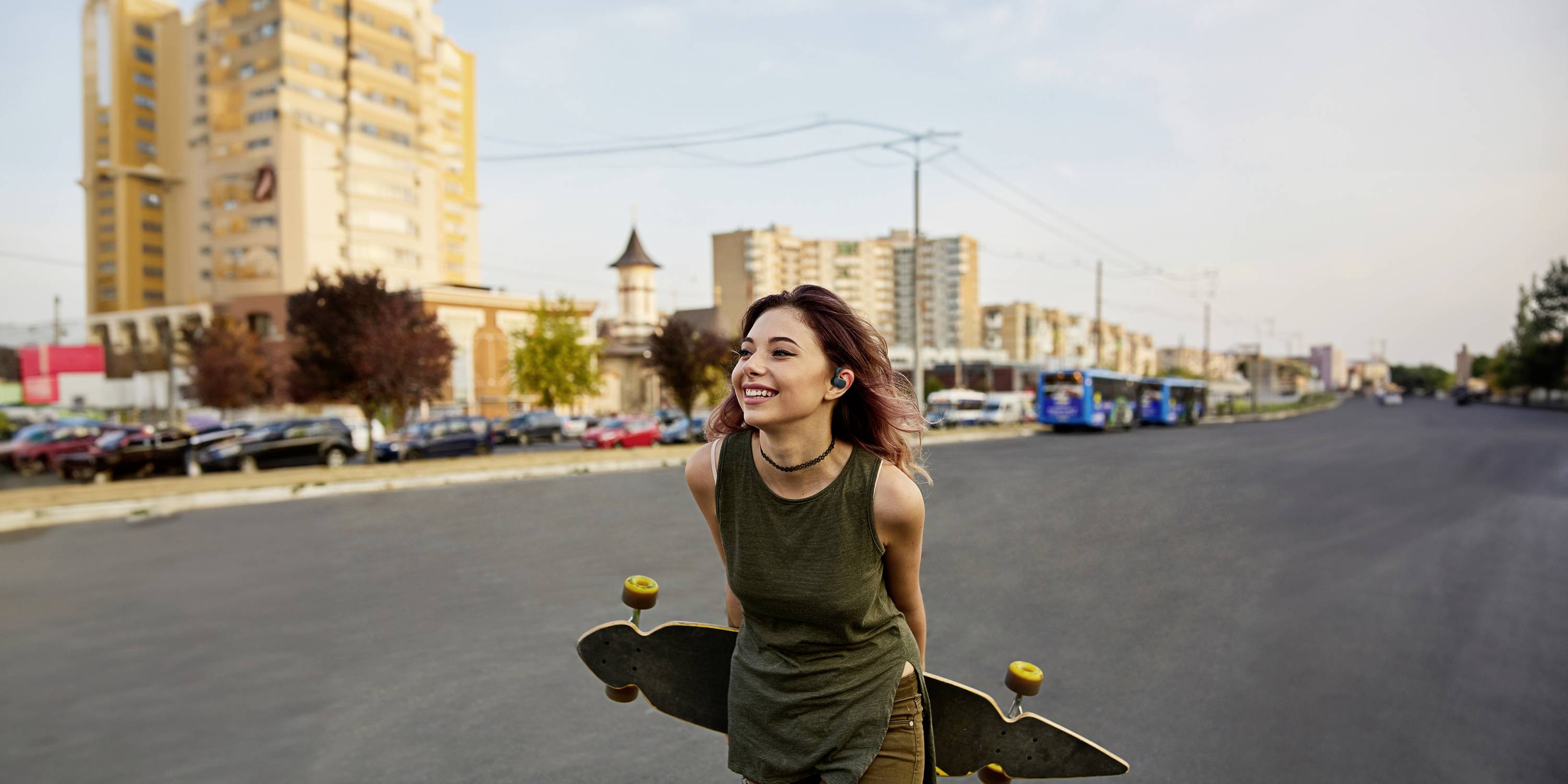 Nasmijana žena drži longboard i trči širokom cestom tijekom sunčanog dana. U pozadini se vide zgrade.