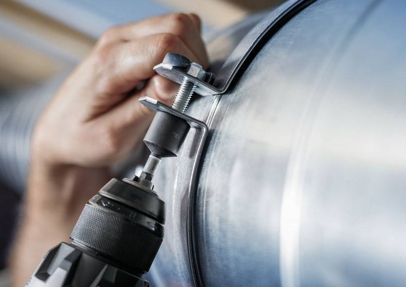 Jedna ruka drži kliješta za cijev koja je pričvršćena za srebrnu cijev, dok bušilica buši rupu u cijev.