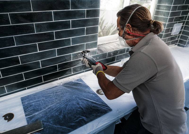 Jedna osoba reže pločice unutra električnim alatom na radnoj ploči. Nosi zaštitne naočale i masku.