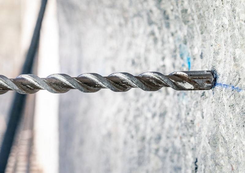 Metalni bušač probija preciznu rupu u sivoj betonskoj stijeni. Fokus je na bušaču u sredini slike.