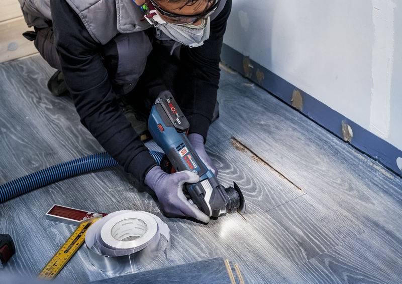 Jedna osoba obavlja popravak na sivom drvenom podu pomoću električne alatke. Vidljiva je zaštitna odjeća i oprema.