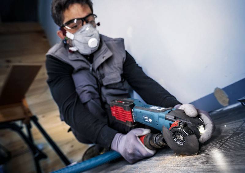 Radnik u zaštitnoj opremi reže drvo električnom brusilicom. Nosi zaštitnu masku za prašinu i zaštitne naočale.