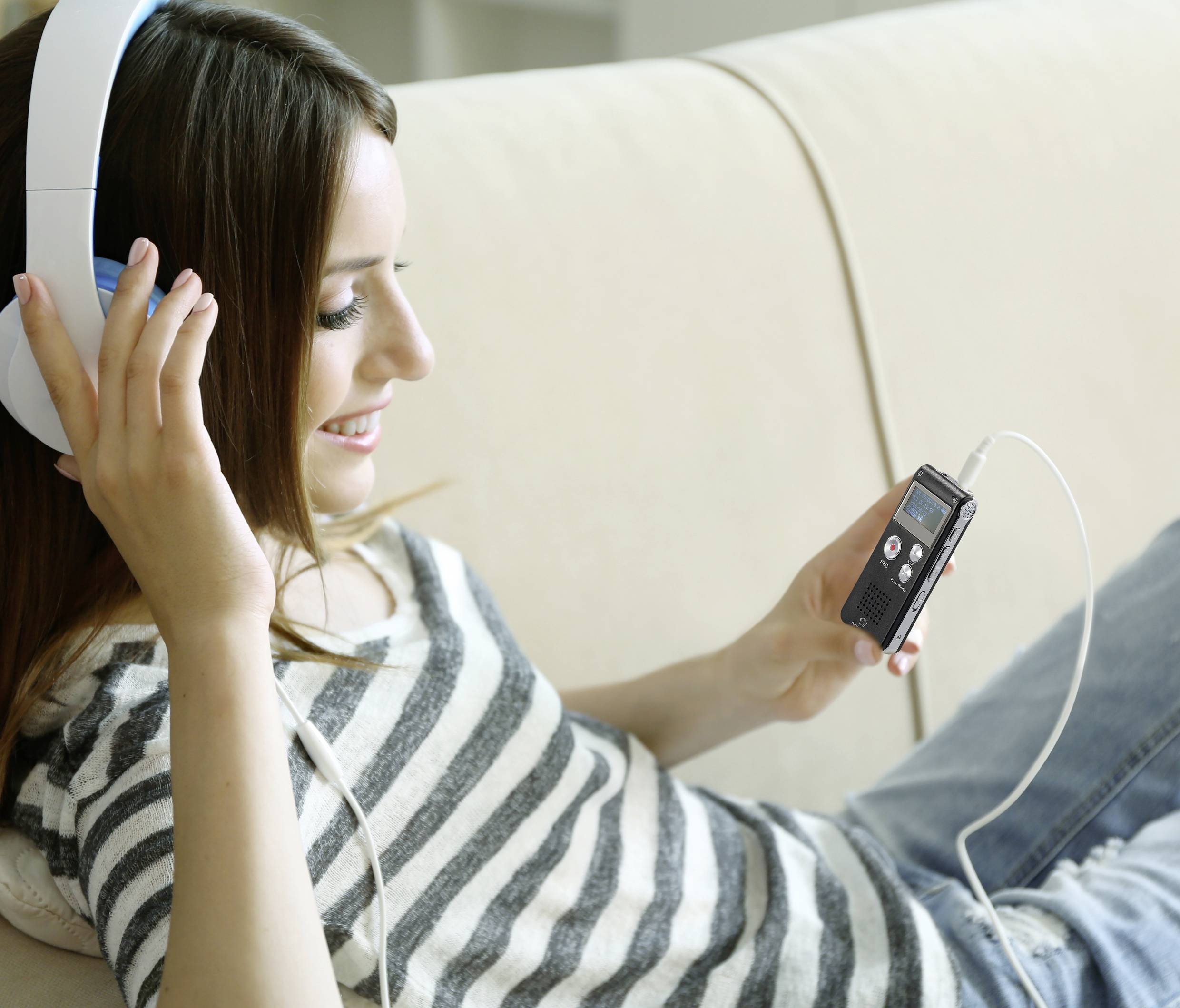 Žena opušteno leži na kauču, sluša glazbu slušalicama i drži prijenosni audio uređaj u ruci.