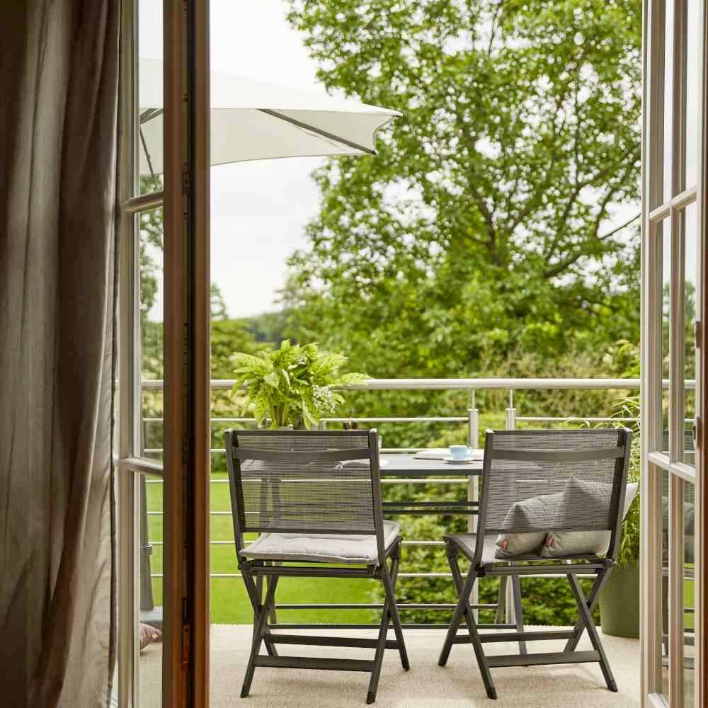 Pogled iznutra na balkon s dva stolca i stolom, okružen zelenim površinama i velikim stablom u pozadini.