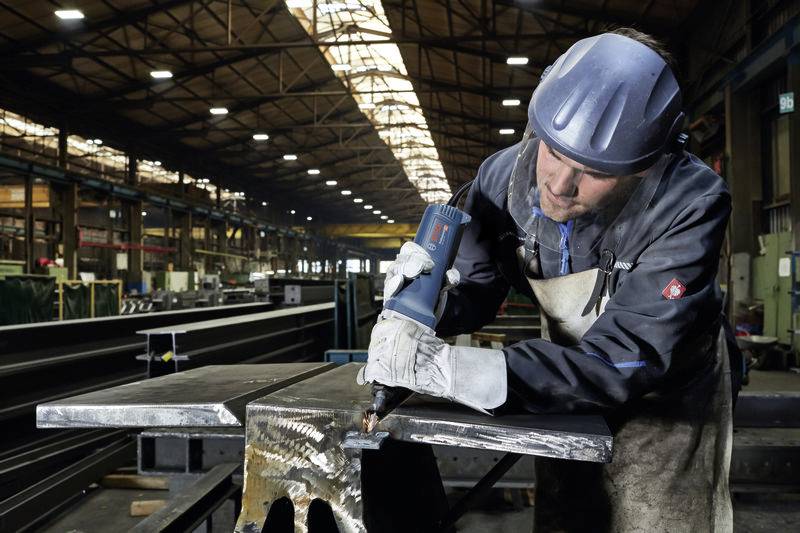 Radnik u zaštitnoj opremi zavaruje metal u velikoj tvorničkoj hali. U pozadini su vidljivi čelični nosači i industrijski strojevi.