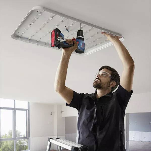 Muškarac na ljestvama montira veliku LED stropnu lampu akumulatorskim odvijačem u svijetloj prostoriji.