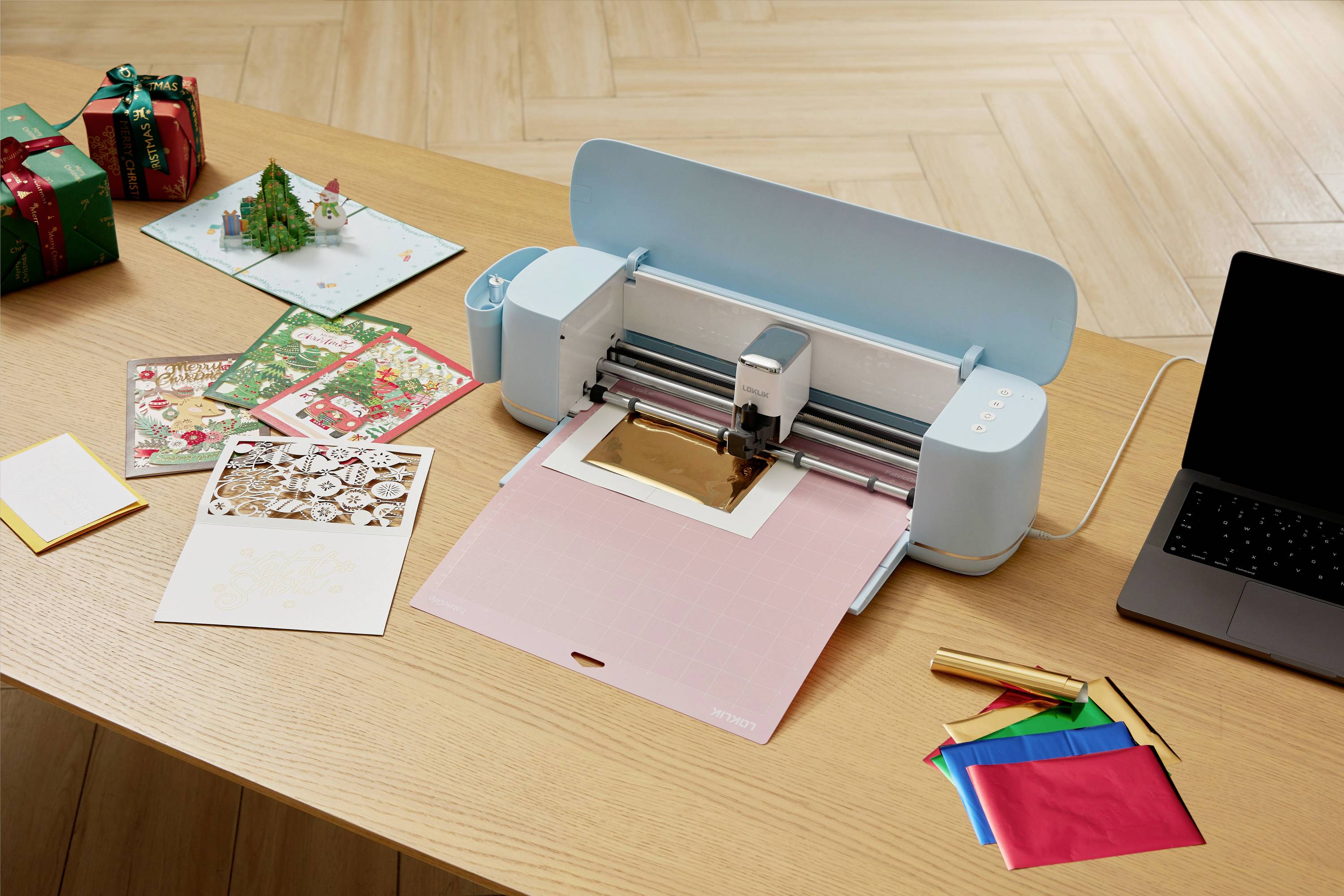 Uređaj za rezanje papira (plotter) na stolu reže papir, okružen kartama, ukrasnim papirom i prijenosnim računalom.