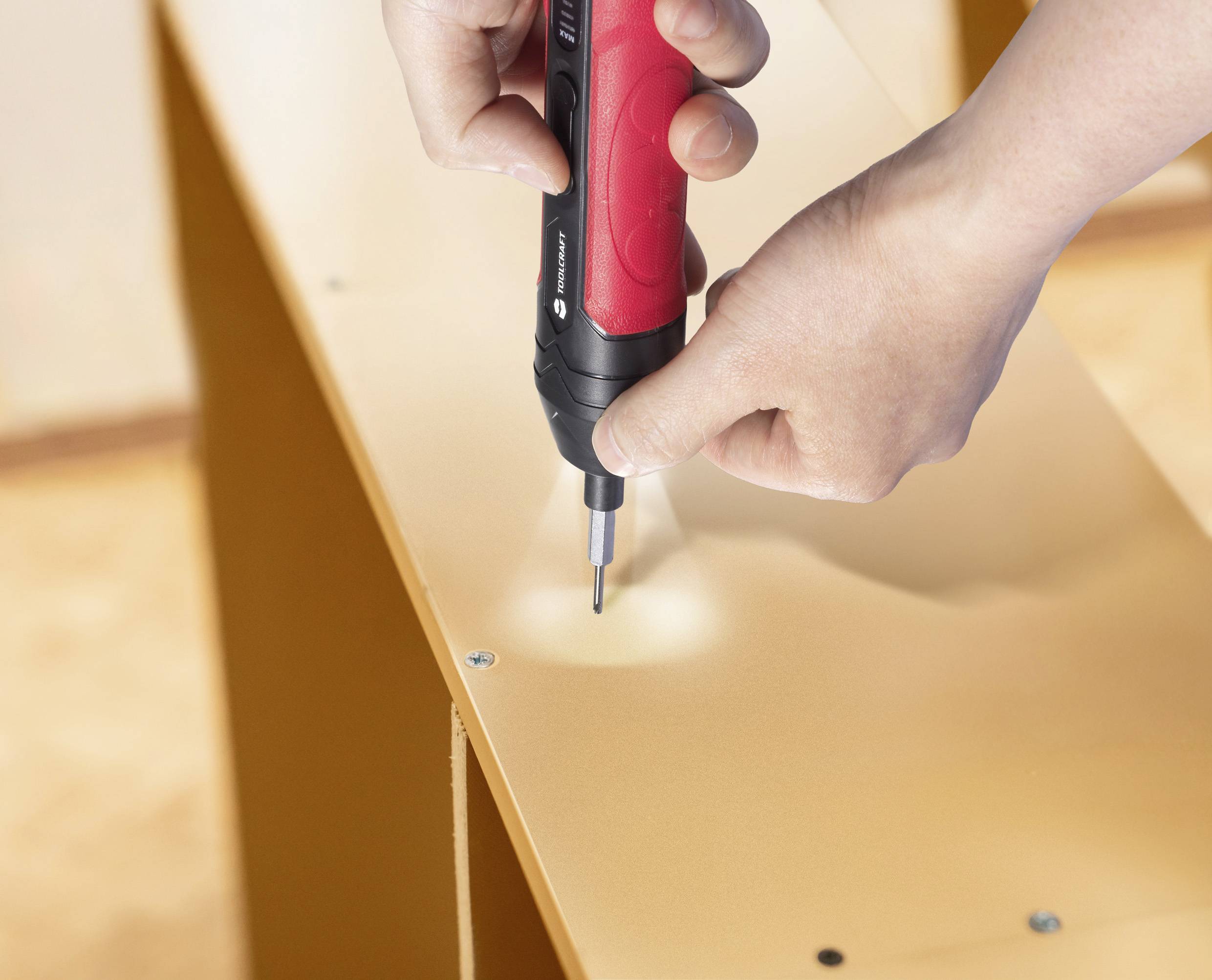 Ruke koje koriste crveni i crni električni odvijač za sastavljanje namještaja, uvrtajući vijke u drvenu površinu.