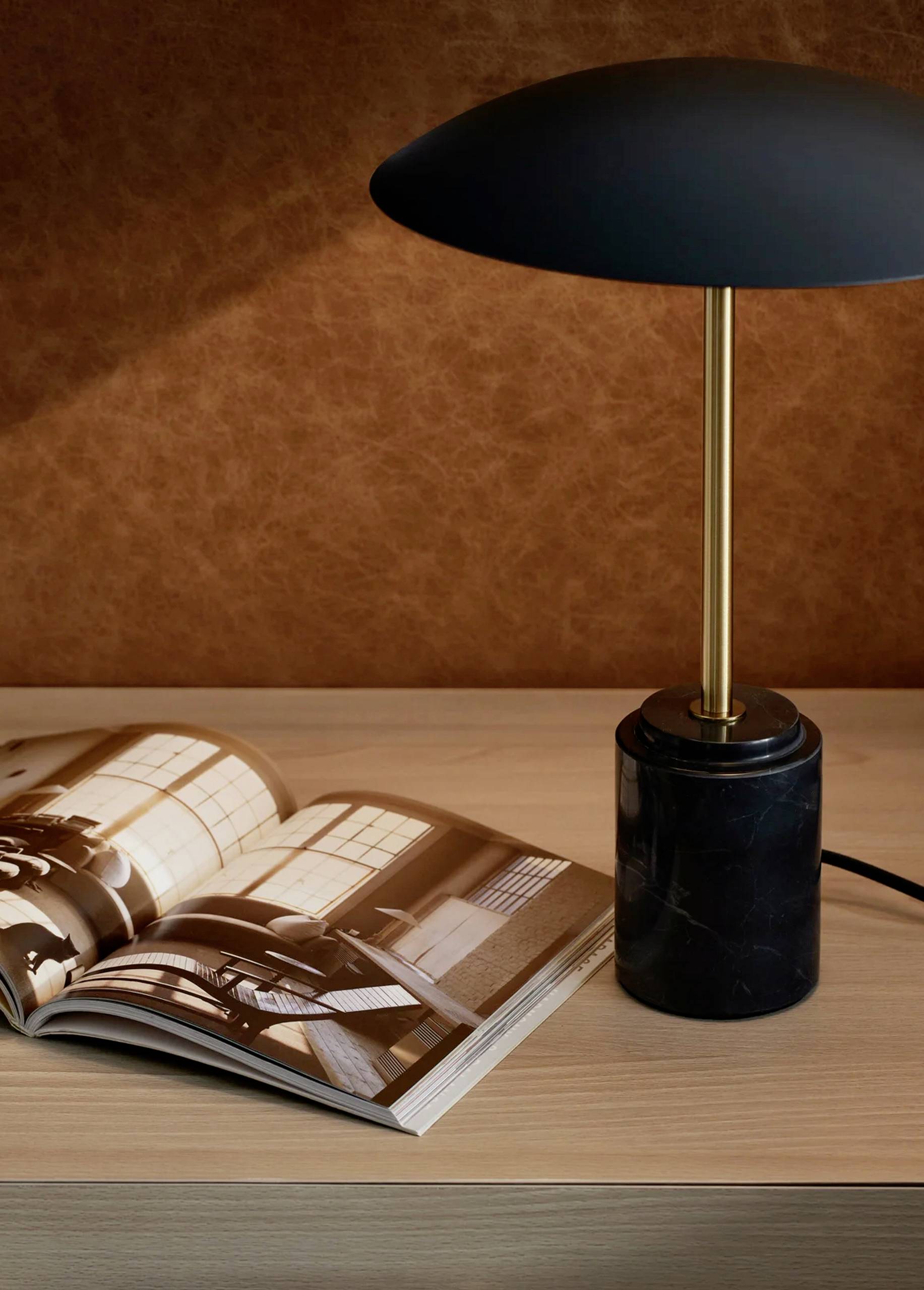 Otvoreni dizajnerski časopis leži na drvenom stolu pored moderne stolne lampe s crnim sjenilom i zlatnim postoljem.