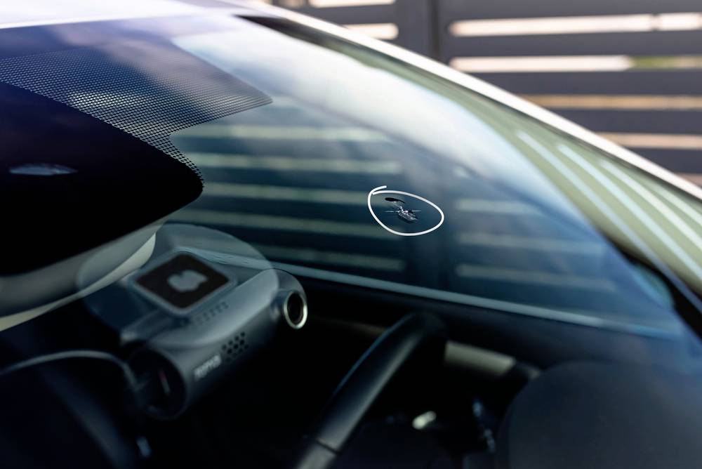 Prednje staklo automobila s kružno označenim udarcem kamena. U pozadini se nejasno naziru rešetke.