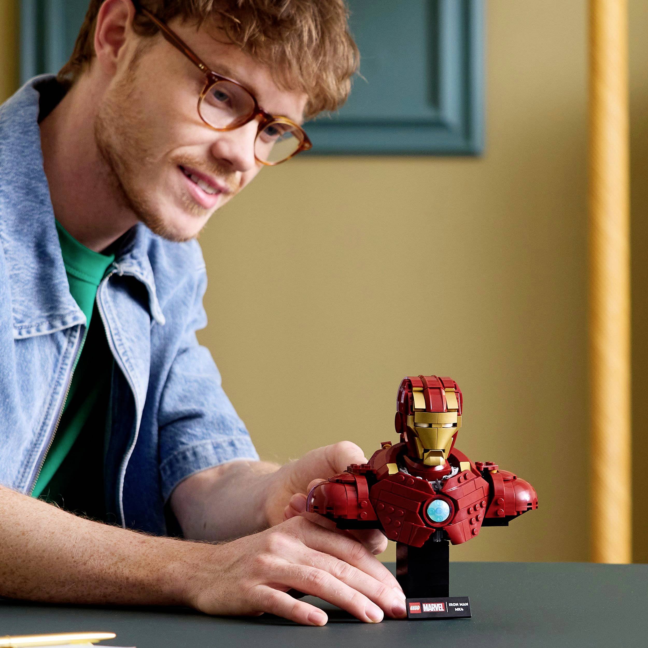 Muškarac s naočalama se smiješi i sjedi za stolom, pred njim stoji Iron Man figura od LEGO kockica s malom pločicom.