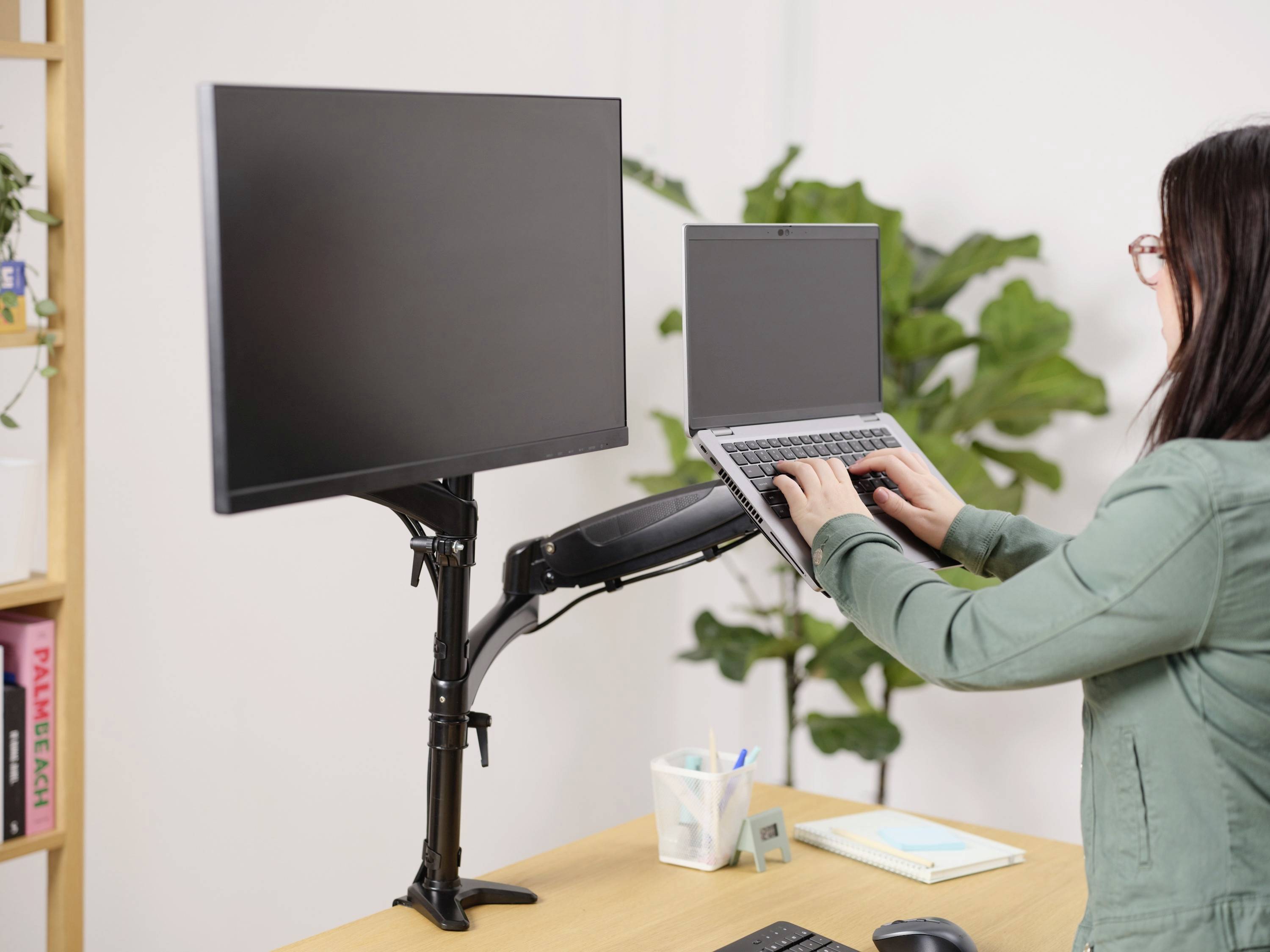 Osoba stoji uz radni stol i koristi laptop i dva monitora na podesivom stalku. U pozadini se nalazi biljka.