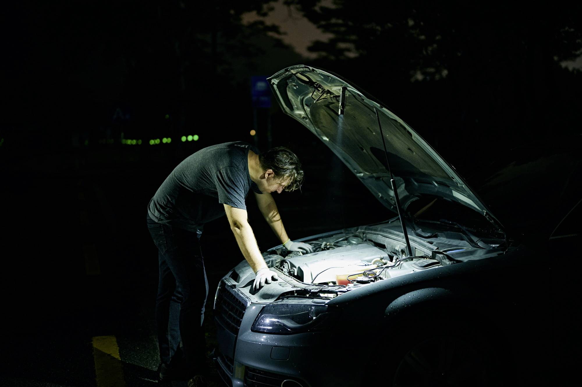 Muškarac stoji noću pored automobila s otvorenim poklopcem motora na mračnoj cesti. Pregledava motor pomoću džepne lampe.