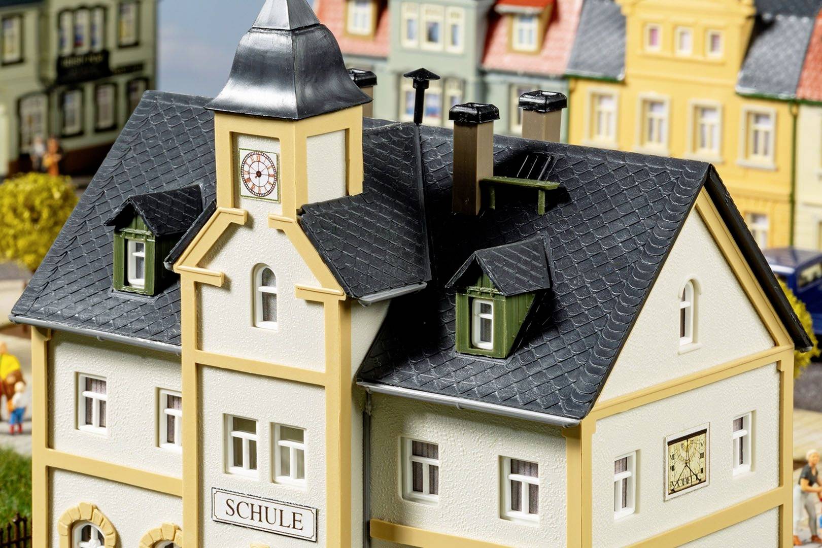 Miniatura škole sa sivim krovom, satnom kulom i okolnim šarenim zgradama u modelnom gradu.