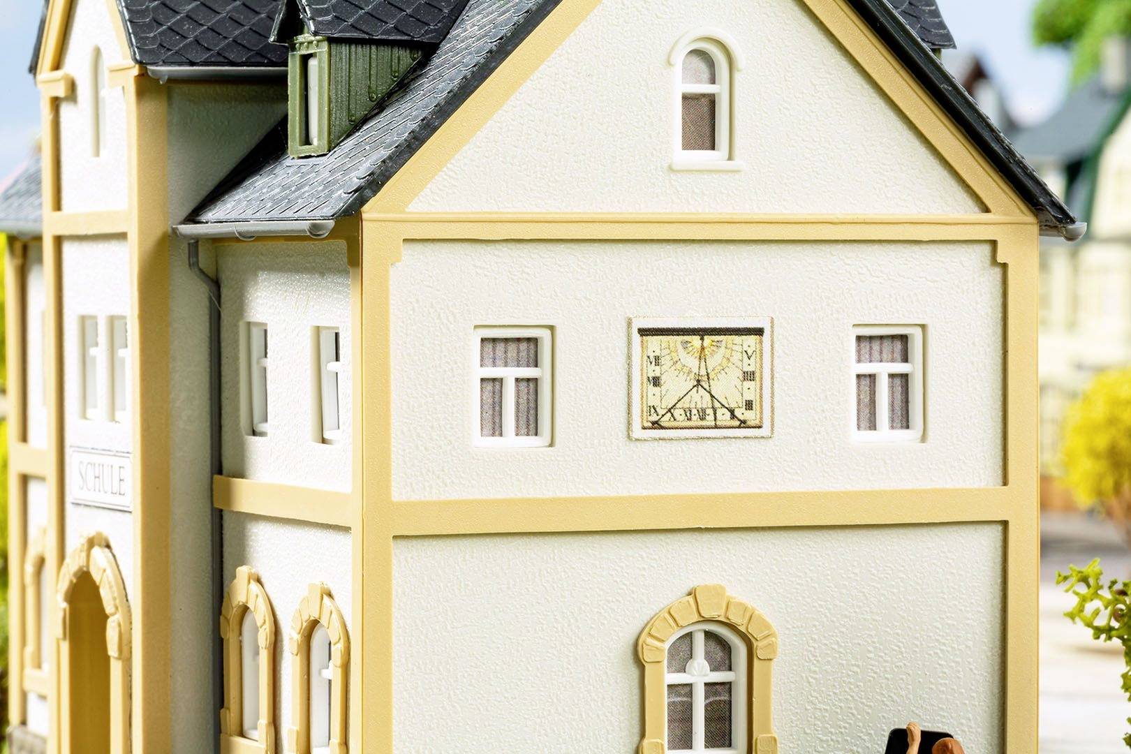 Model zgrade s bež zidovima i sivim krovom prikazuje sat na prednjoj strani. U pozadini su vidljive dodatne modelske strukture.