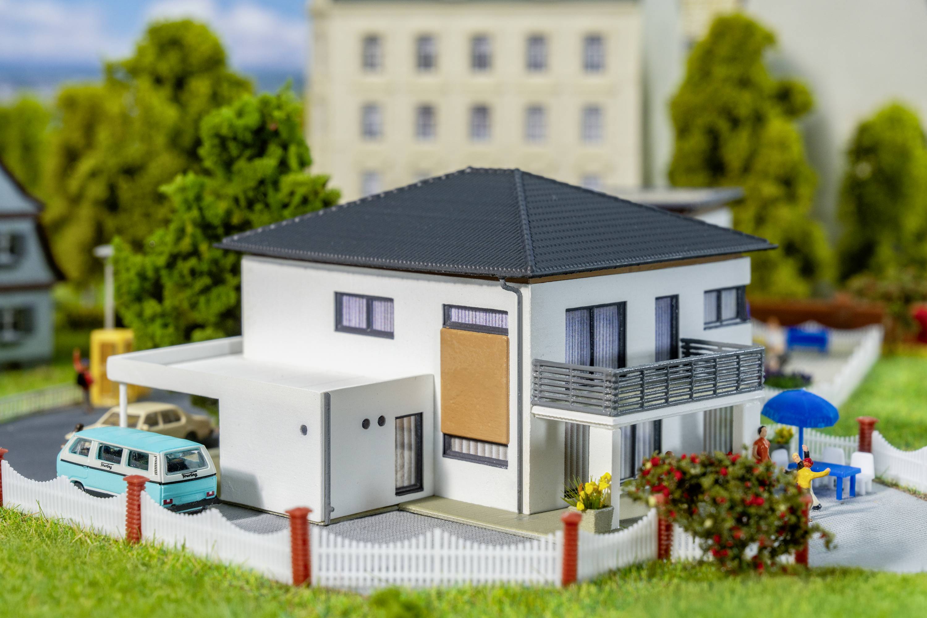Moderna obiteljska kuća s ravnim krovom i balkonom nalazi se u zelenom okruženju, s plavim automobilom i suncobrano u vrtu.