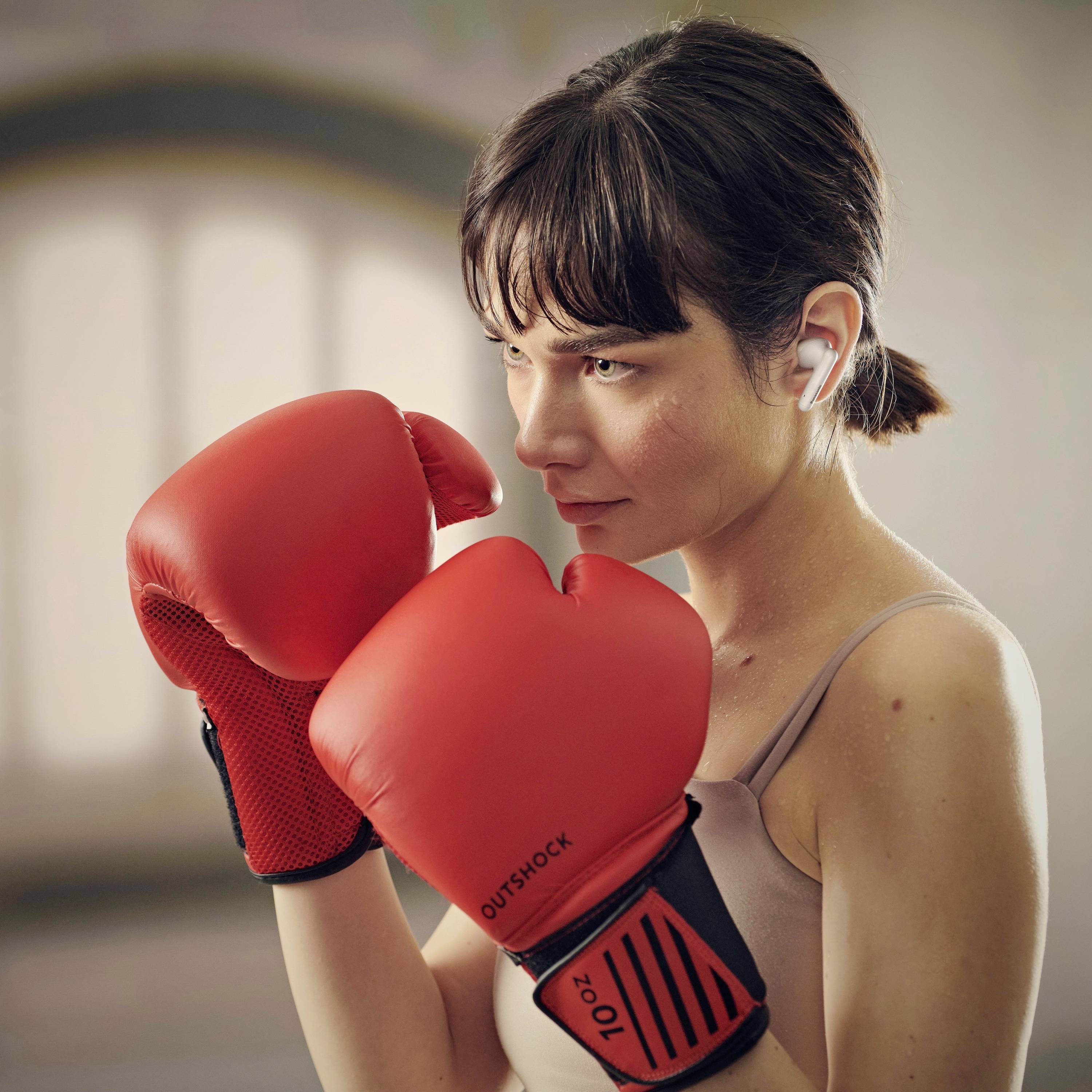 Žena u boksačkim rukavicama stoji u borbenoj poziciji, intenzivno usredotočena. Nosi bežične slušalice, što ukazuje na fitness ili trening.