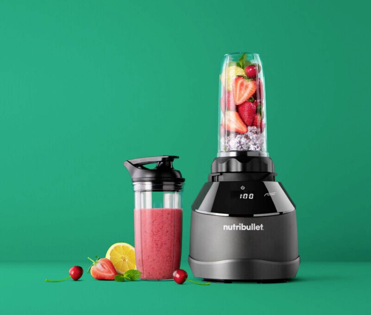 Blender pun voća pokraj smoothieja u boci, postavljen na zelenu pozadinu. Vidljiv je brend 'Nutribullet'.