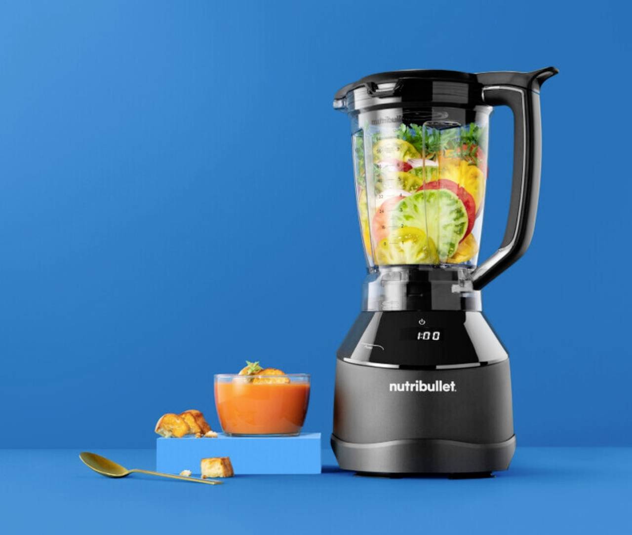 Blender napunjen različitim voćem i povrćem pokraj zdjele juhe i žlice, postavljeni na svijetloj plavoj pozadini.
