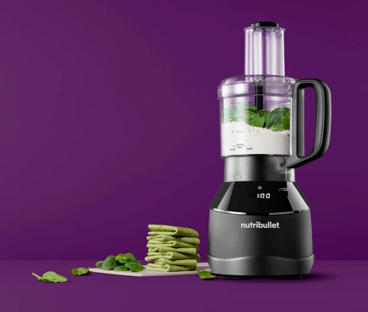 Blender na ljubičastoj pozadini koji miješa sastojke, s komadićima zelenih palačinki i špinatom na stolu u blizini.