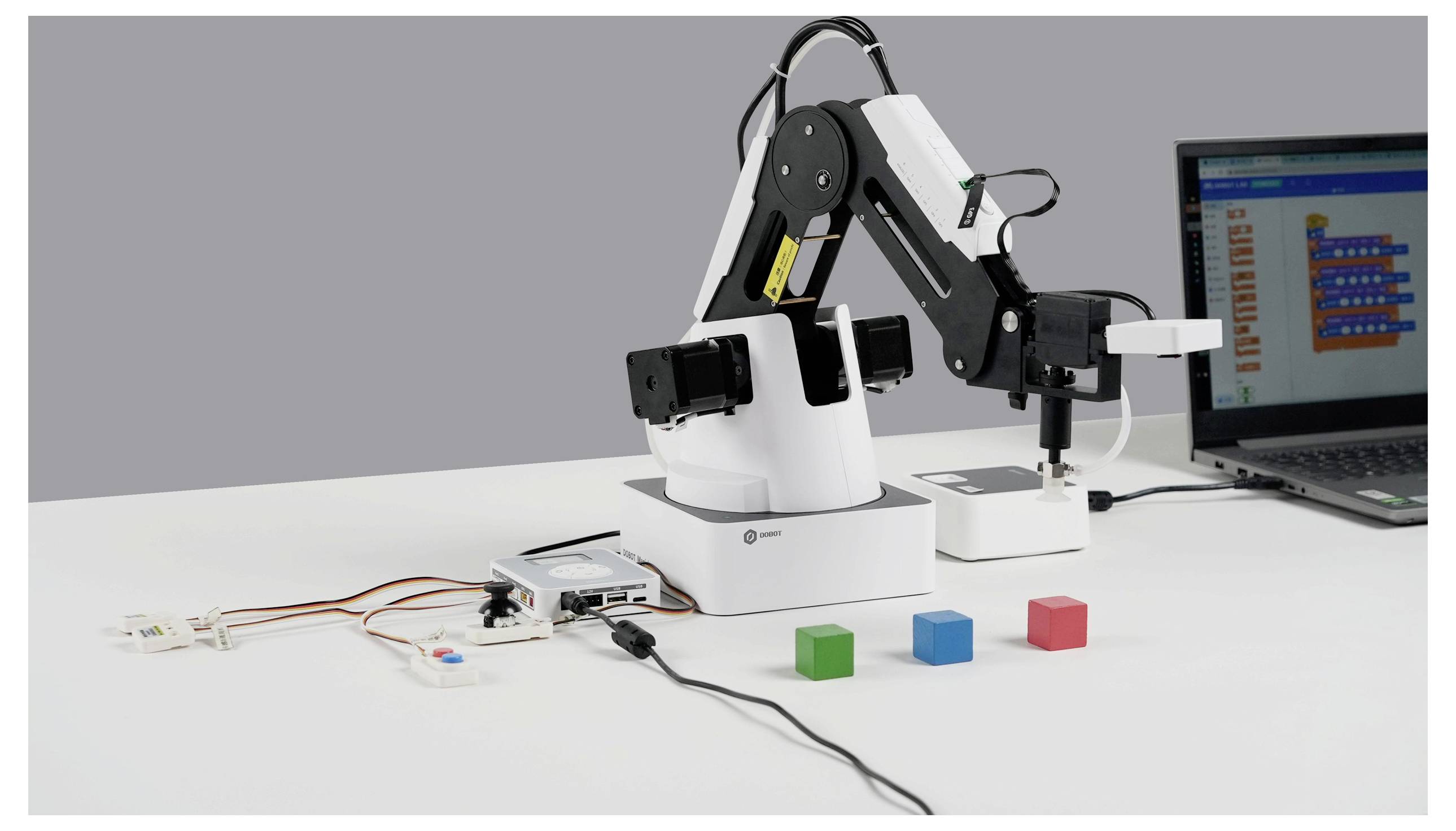 Robotska ruka na radnom stolu komunicira s obojenim blokovima pored prijenosnog računala koje pokće softver za programiranje, prikazujući učenje robotike.
