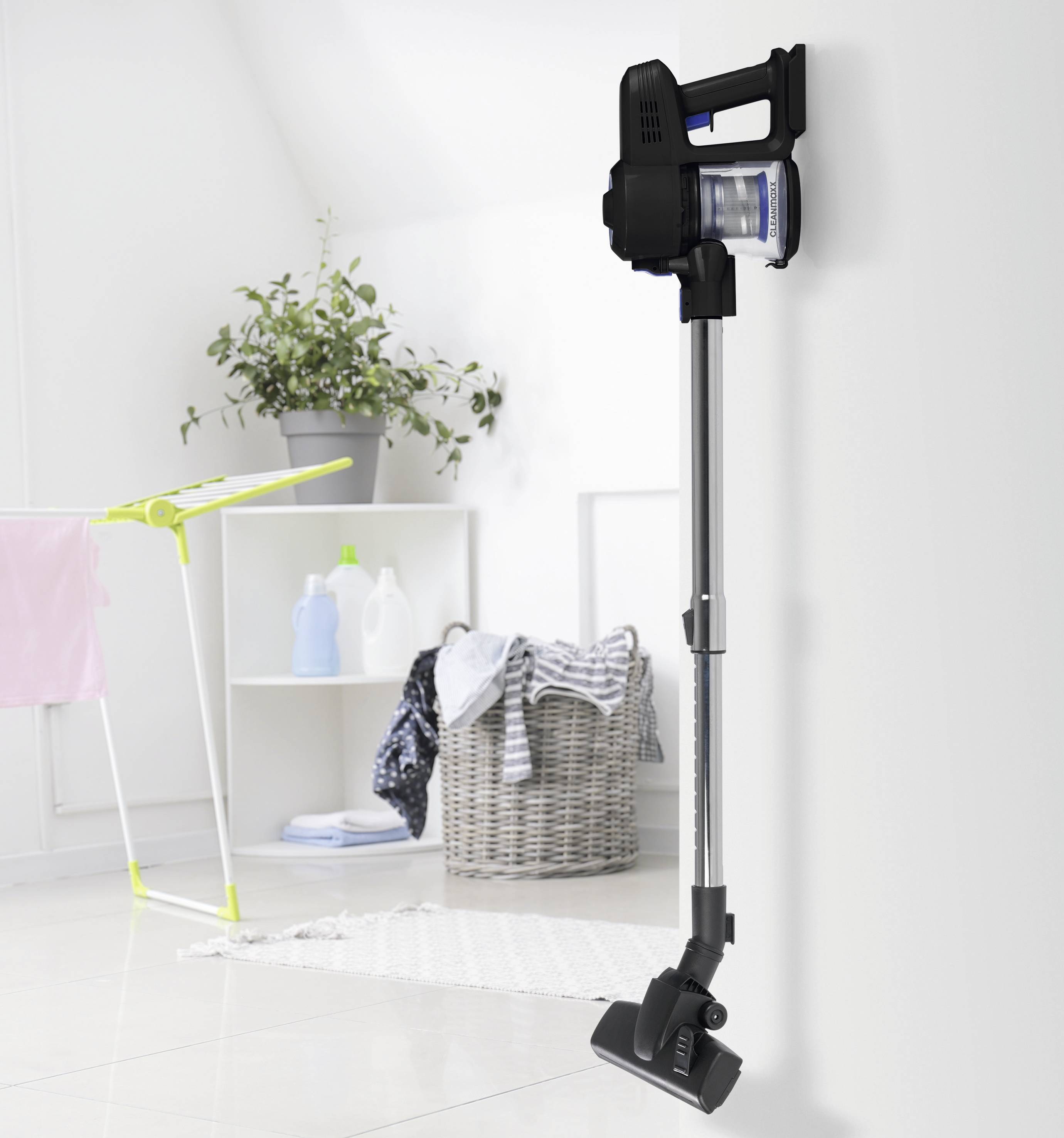 Bežični usisavač postavljen na zid u svijetlom, organiziranom prostoru za dnevni boravak s košarom za rublje, sušačem rublja i ukrasnim cvijetom u posudi u blizini.