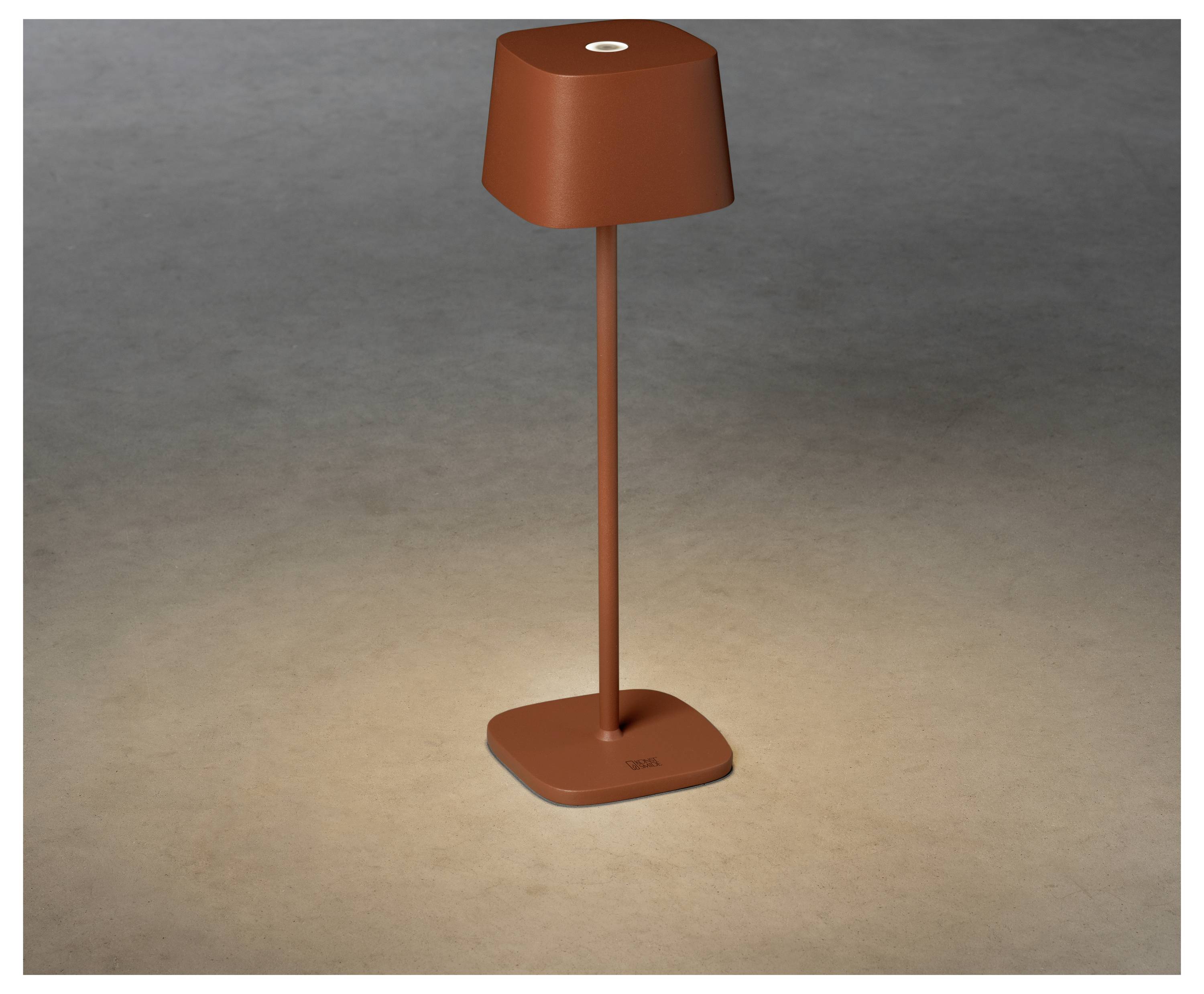 Elegantan, moderan stolni lampa s kvadratnom bazom u crvenosmeđoj boji, koja osvjetljava površinu ispod nje blagim, toplim svjetlom.