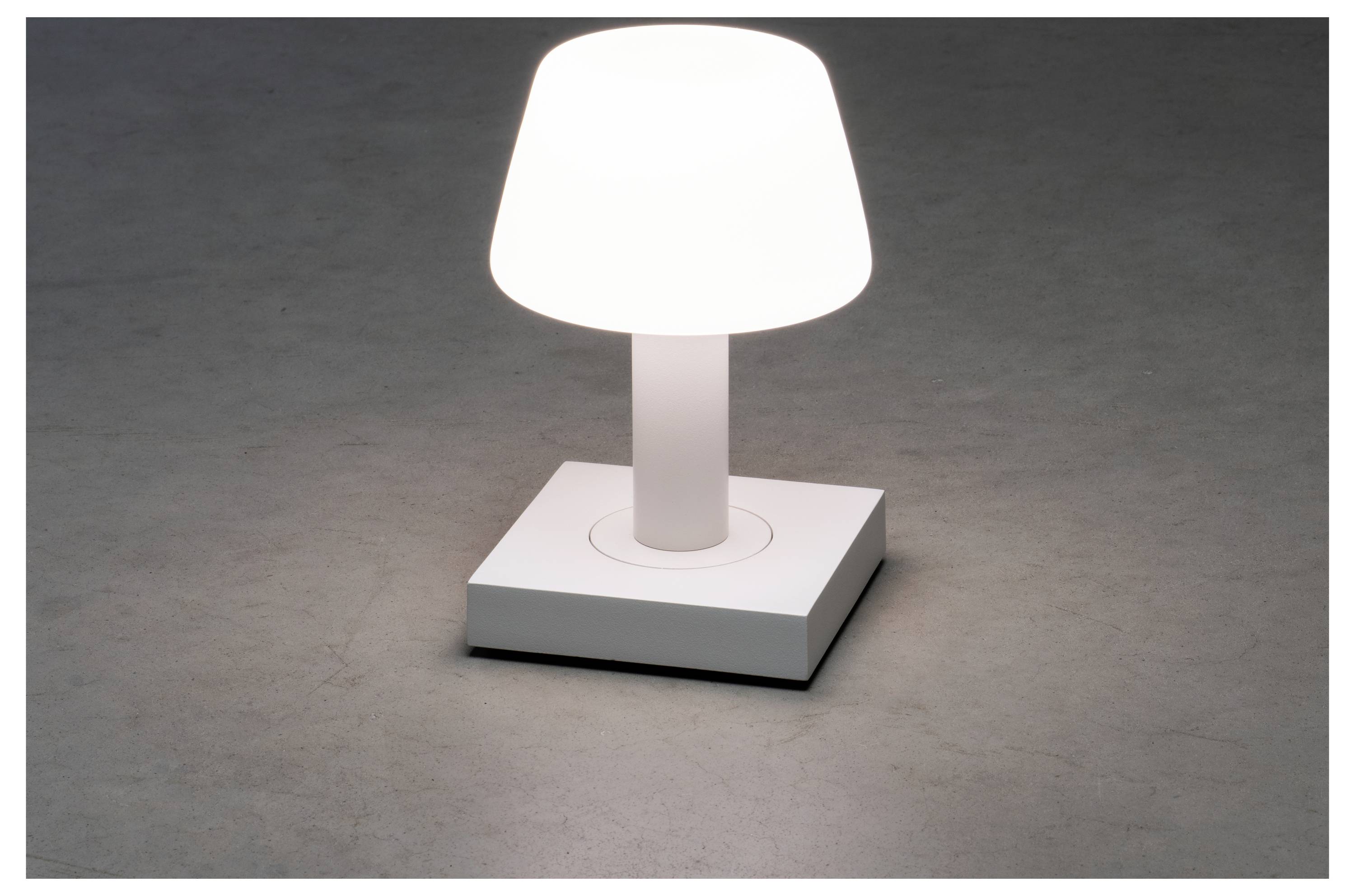 Mala bijela stolna lampa kvadratnog postolja emitira meko svjetlo na sivoj površini, stvarajući minimalistički i moderan estetski dojam.
