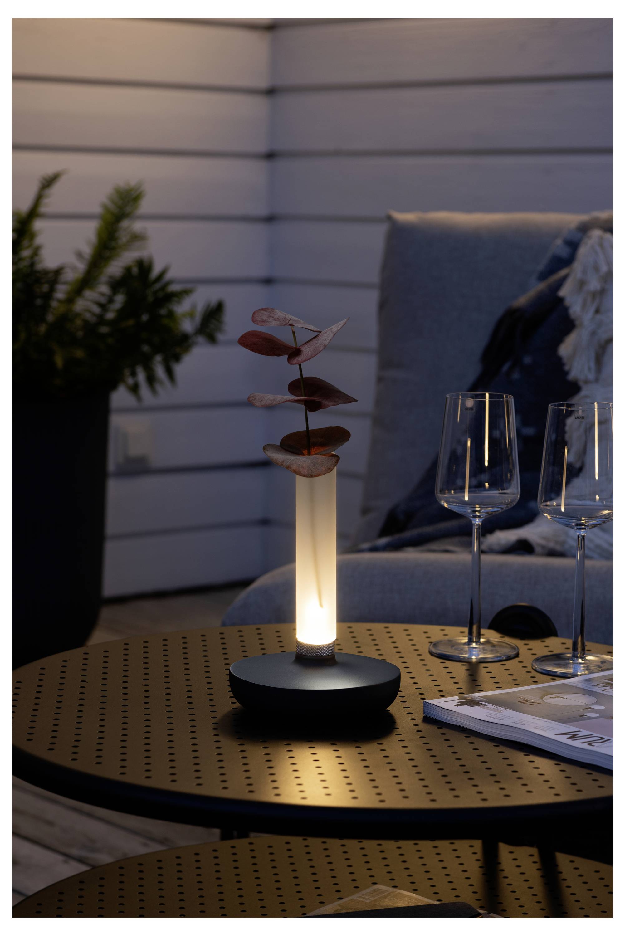 Moderna stolna lampa emitira toplo svjetlo na terasi, pored dvije prazne čaše za vino i magazina, uz biljku u pozadini.