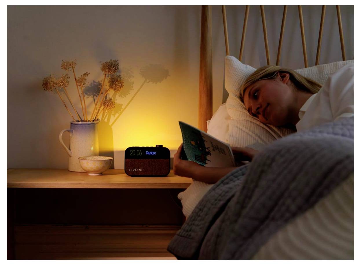 Osoba leži u krevetu čitajući knjigu pokraj tople lampe. Sat za alarm prikazuje '20:06 Opusti se'. Okruženje je mirno i ugodno.