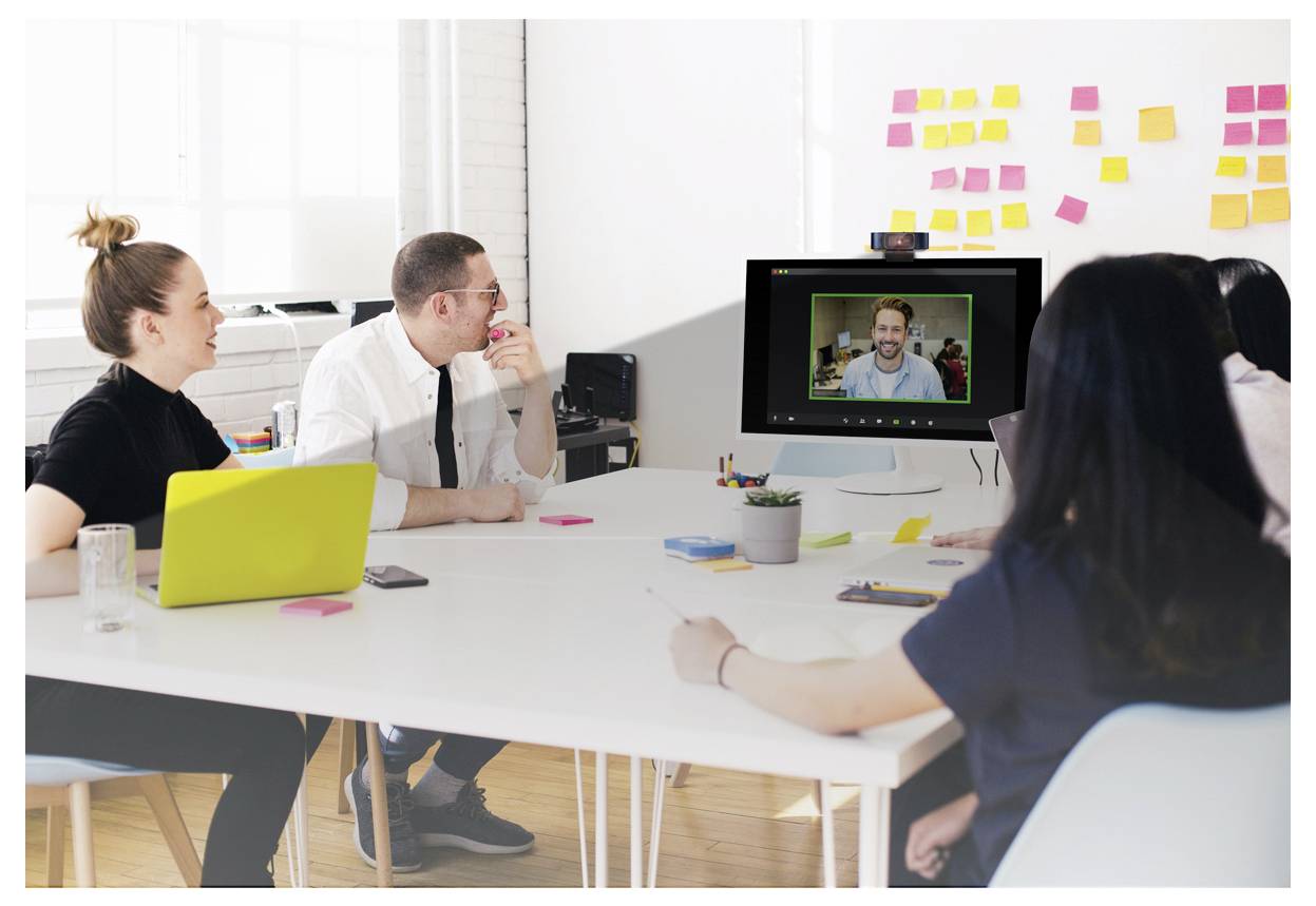 Grupa od četiri osobe u prostoriji za sastanke sudjeluje u video pozivu s osobom prikazanom na monitoru. Svijetla prostorija s lijepljivim bilješkama.