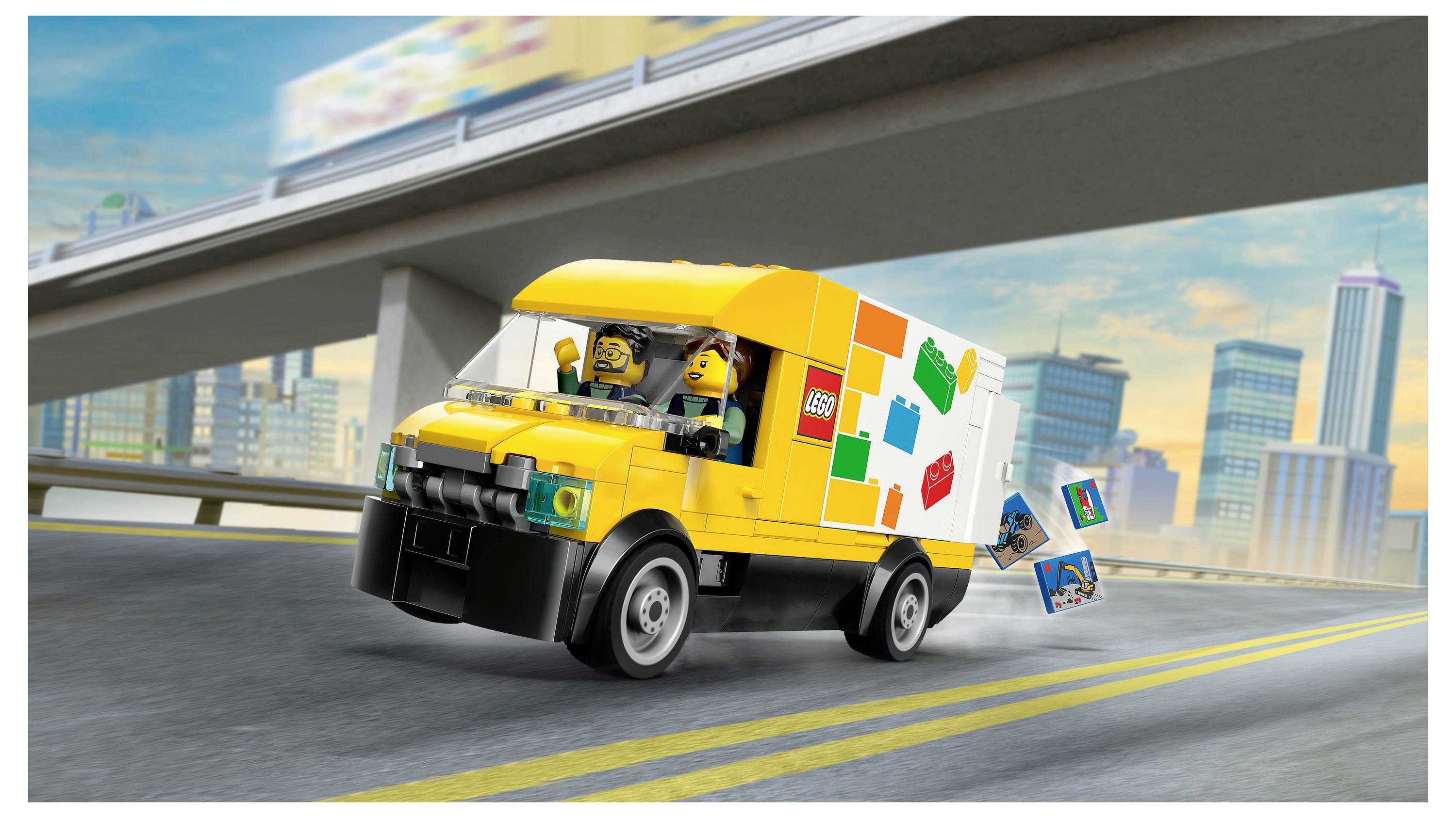 Žuti igračka dostavni kamion koji vozi autocestom s LEGO figuricama unutra, s kulisama grada u pozadini. Kamion ima LEGO kockice na svojoj bočnoj stranici.