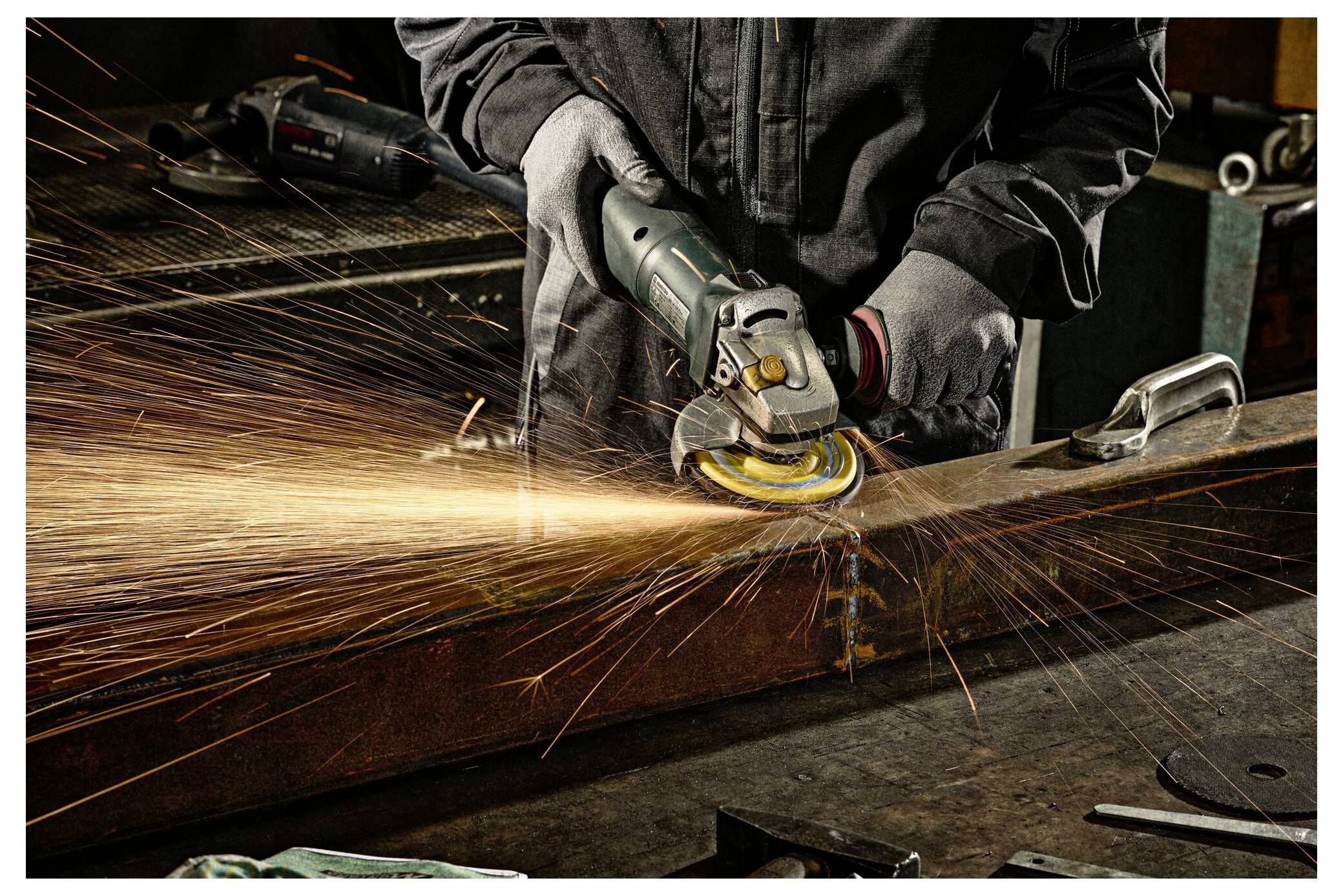 Osoba u zaštitnoj opremi koristi ugaonu brusilicu za rezanje metala, stvarajući sjajne varnice u radionici. Fokus je na alatu i radnji.