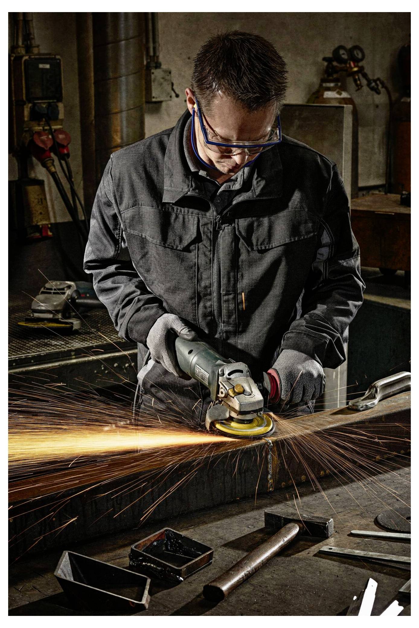 Osoba u sigurnosnoj opremi koristi brusilicu na metalnoj površini u radionici, stvarajući svijetle narančaste iskre. U pozadini se nalaze alati i oprema.