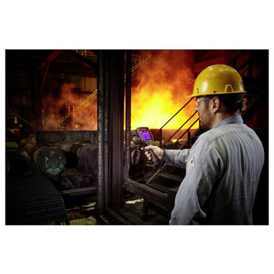 Radnik u zaštitnoj opremi koristi termičku kameru u blizini žarko narančaste peći unutar industrijske postrojenja.