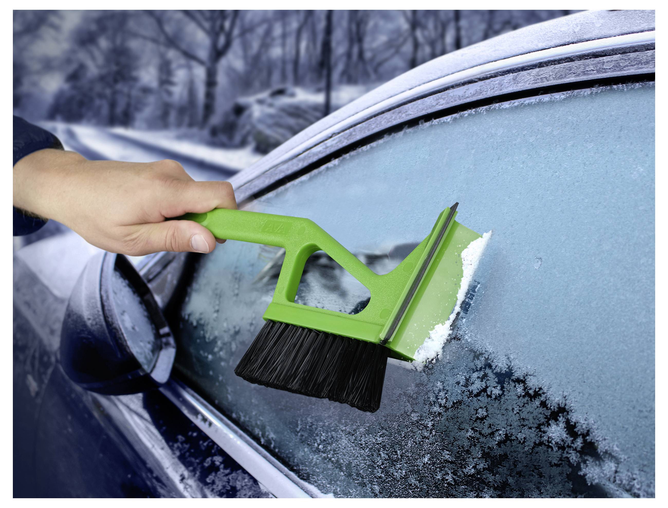 Ruka koristi zeleni strugač leda da ukloni mraz s prozora automobila na snježnoj zimskoj cesti.