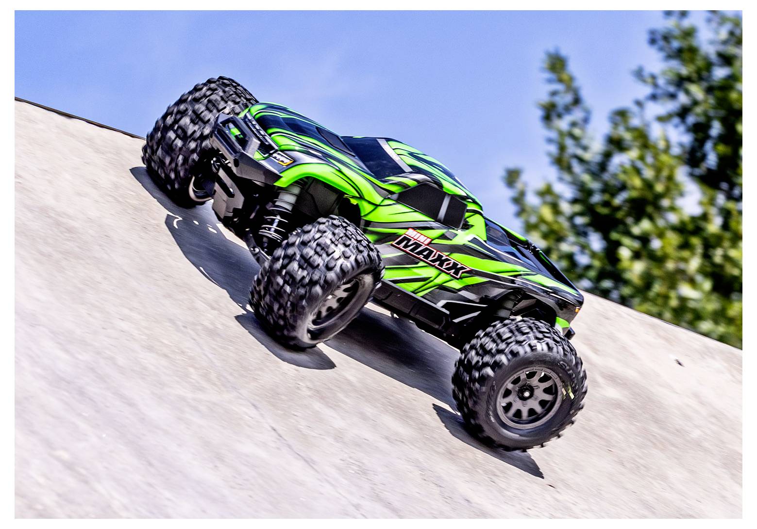 Zeleni i crni daljinski upravljani monster truck vozi uzbrdo po strmom betonskom nagibu. Stabla su zamućena u pozadini.